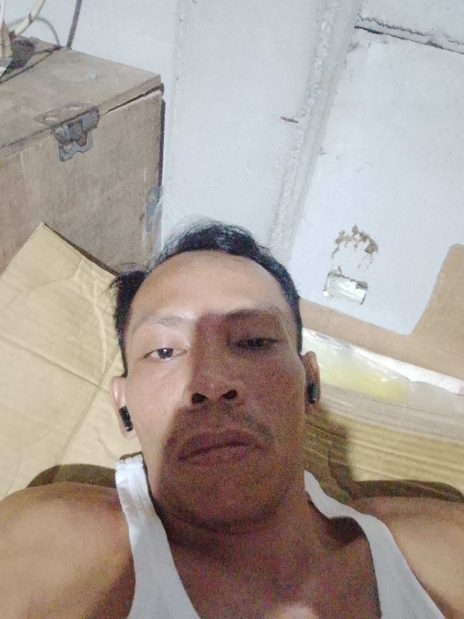 The image presents a medium close-up of an adult male in Bukit Ketok, Indonesia. He is wearing a white tank top and what appears to be a black wireless earbud in his left ear. The background includes a corrugated cardboard box and a painted wall with imperfections. An indistinct wooden box sits in the upper left corner. The lighting is relatively dim, suggesting possible indoor conditions.