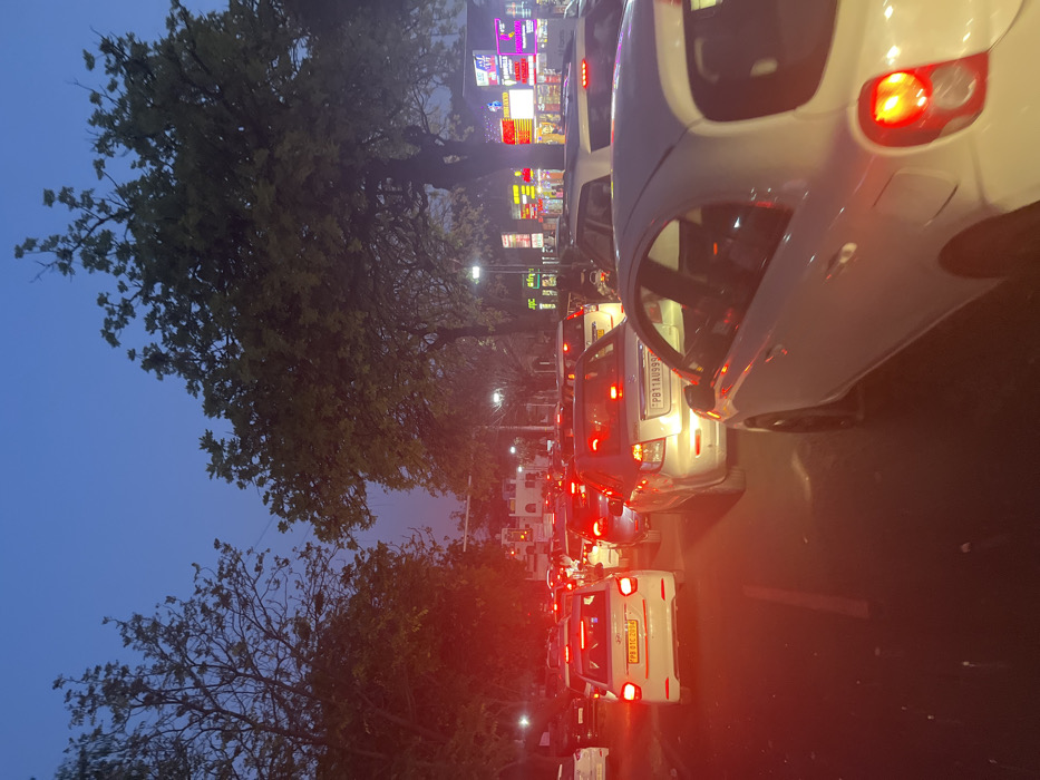 The image shows a traffic jam in Sahibzada Ajit Singh Nagar, India, at 30.715 latitude and 76.718 longitude. The scene is a road lined with trees on either side, and several cars are stopped in the middle of the road. The sky is dark blue and the road is lit by the headlights of the cars. The cars are all facing forward, and their tail lights are visible. There are no people visible. The cars are all standard-sized, and they are all in good condition. The road is paved with asphalt. The traffic appears to be congested with many cars packed together, and the scene is illuminated by the streetlights and the cars' headlights. The environment is urban with shops on the side of the street.