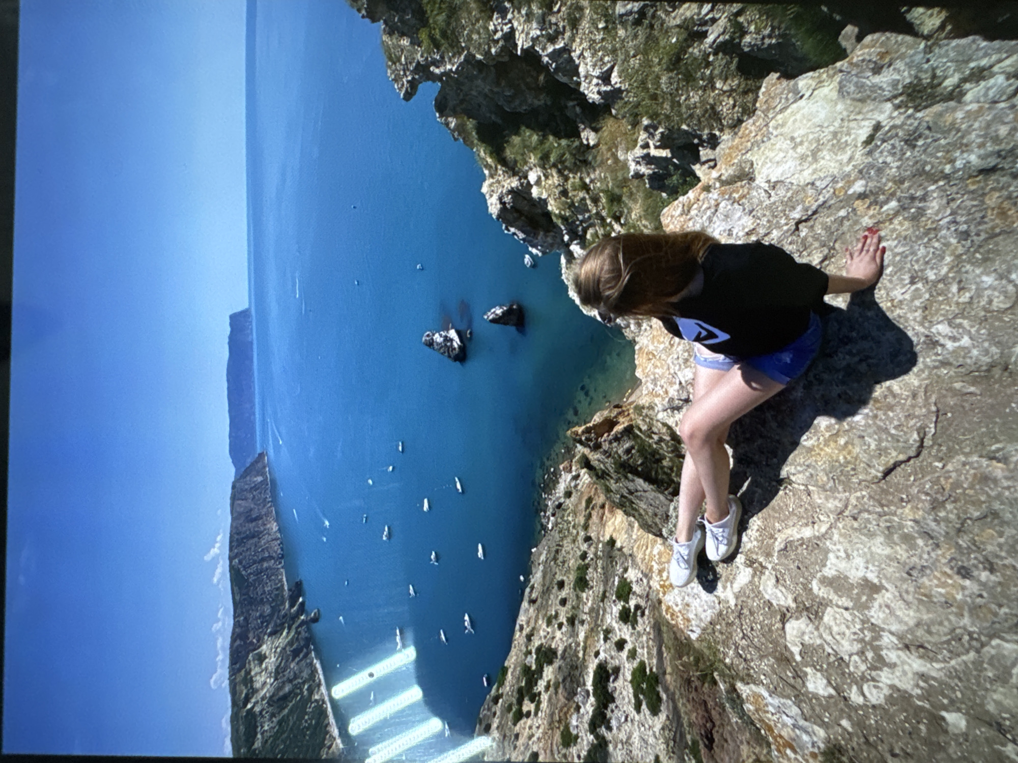 A full shot shows a young woman sitting on a cliff overlooking a bright blue sea with a rocky coastline and numerous boats. The woman is facing away from the camera, looking out at the expansive view. She has long, light brown hair and is wearing a black t-shirt with a white graphic, blue shorts, and white sneakers. Her legs are crossed at the ankle, and she is resting her left arm on the rock beside her, her hand with red nail polish visible. The cliff she is sitting on is a rugged, rocky terrain with sparse green vegetation. The sea is calm and dotted with various boats, including sailboats and motorboats. In the distance, the coastline curves around, with more cliffs and a distant landmass under a clear blue sky. The overall scene is one of natural beauty and peaceful contemplation.