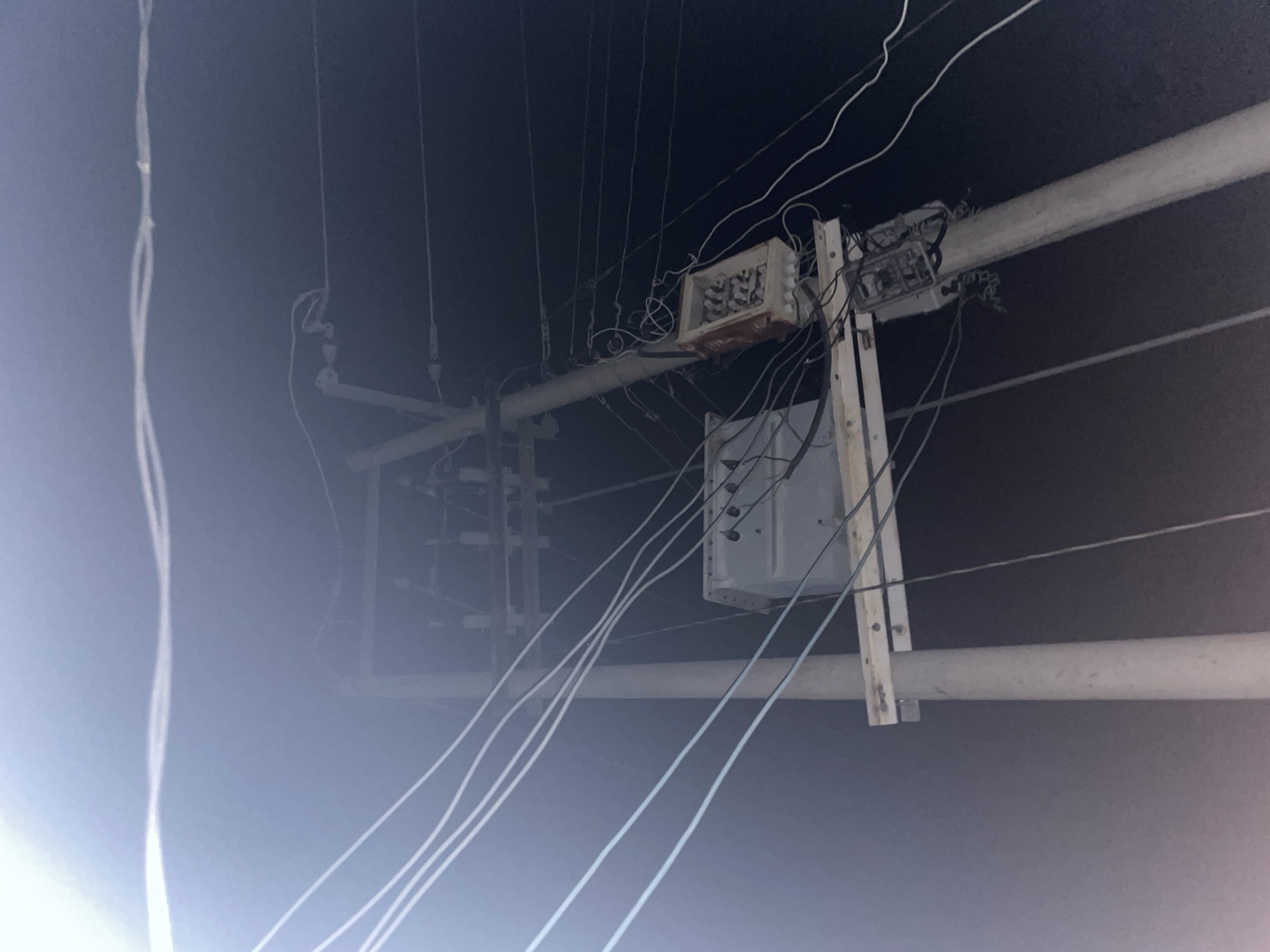 The image shows a utility pole with electrical equipment, likely in Jalingo, Nigeria. The pole is made of concrete and supports a transformer and numerous electrical wires. A junction box, appearing weathered and possibly containing fuses or other electrical components, is mounted to the pole. The background is dark, suggesting nighttime or a dimly lit environment, with a faint glow that could be from streetlights or ambient light. The electrical infrastructure appears aged and somewhat disorganized, with wires strung in various directions. There are no people or animals visible in the scene.