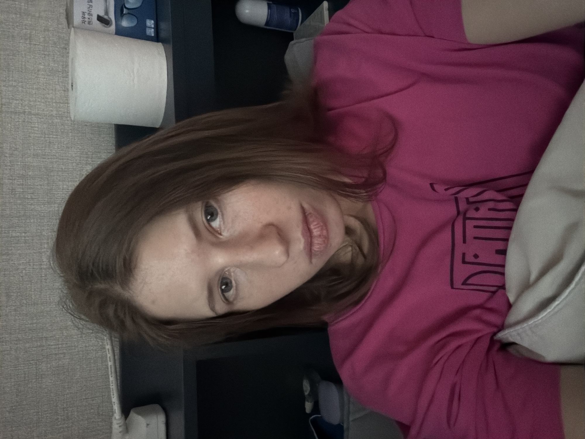 A woman with shoulder-length brown hair is looking at the camera, with her head slightly tilted. She is wearing a bright pink t-shirt with a graphic design. She is lying down, with a pillow supporting her head and upper body. Behind her, on a dark shelf, are various objects, including a roll of toilet paper and a blue box with Korean text and an image of glasses. To the left of the woman, part of a white electrical device is visible. The background appears to be a wall with a textured grey surface. The scene is indoors, likely in a residential setting in Dongducheon-si, South Korea.