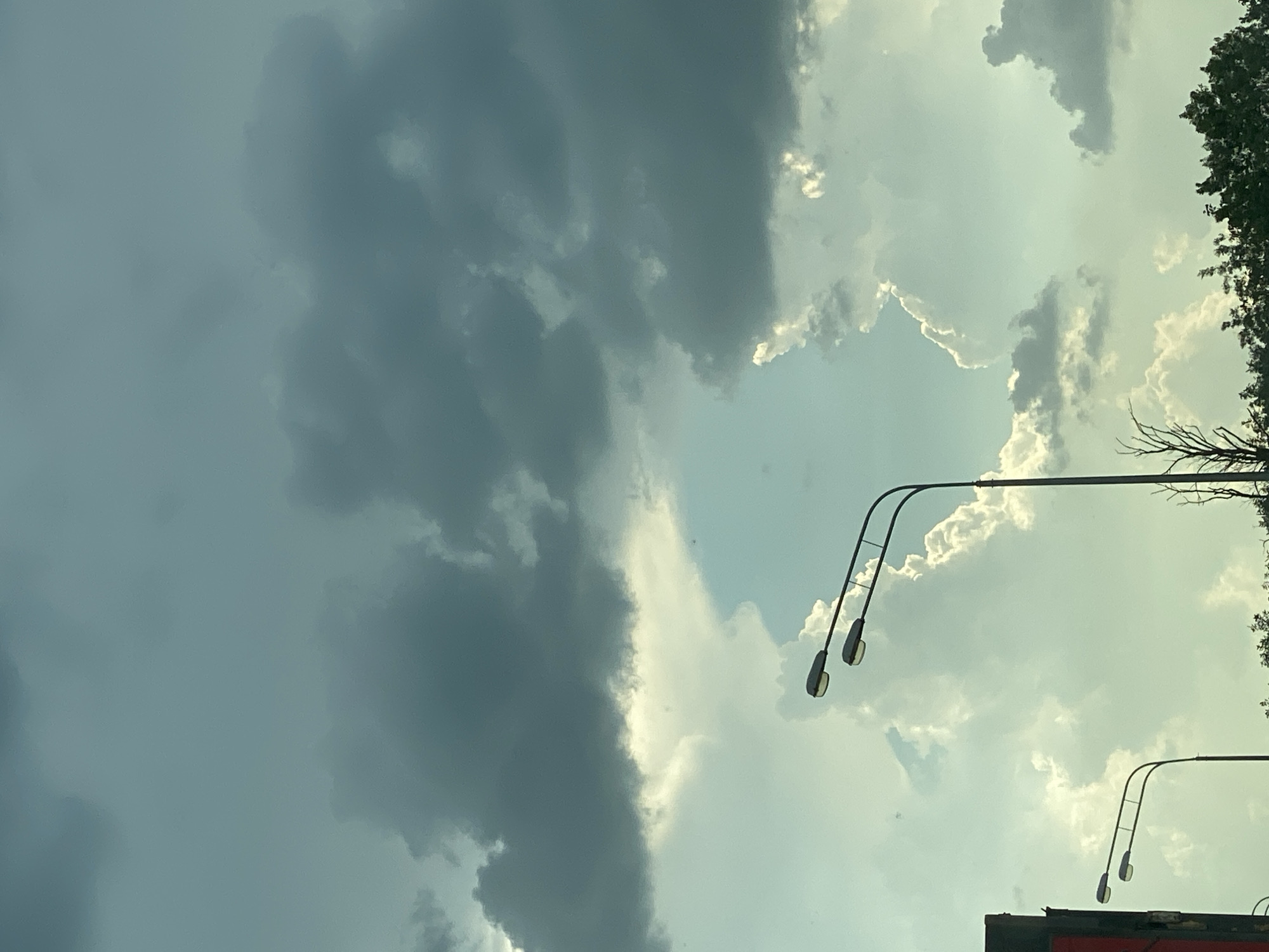The image shows a cloudy sky with two streetlights visible in the foreground. The sky is a mixture of dark grey and lighter, bright white clouds, suggesting a dynamic weather system. The streetlights are modern, with multiple lamps on each pole, and they are positioned on the left and right sides of the frame, partially obscuring the sky. Below the streetlights, a line of green trees and foliage can be seen, with a hint of a red structure on the far left edge of the image. The overall impression is one of an outdoor scene, possibly on a street or highway, under an overcast or transitioning sky.