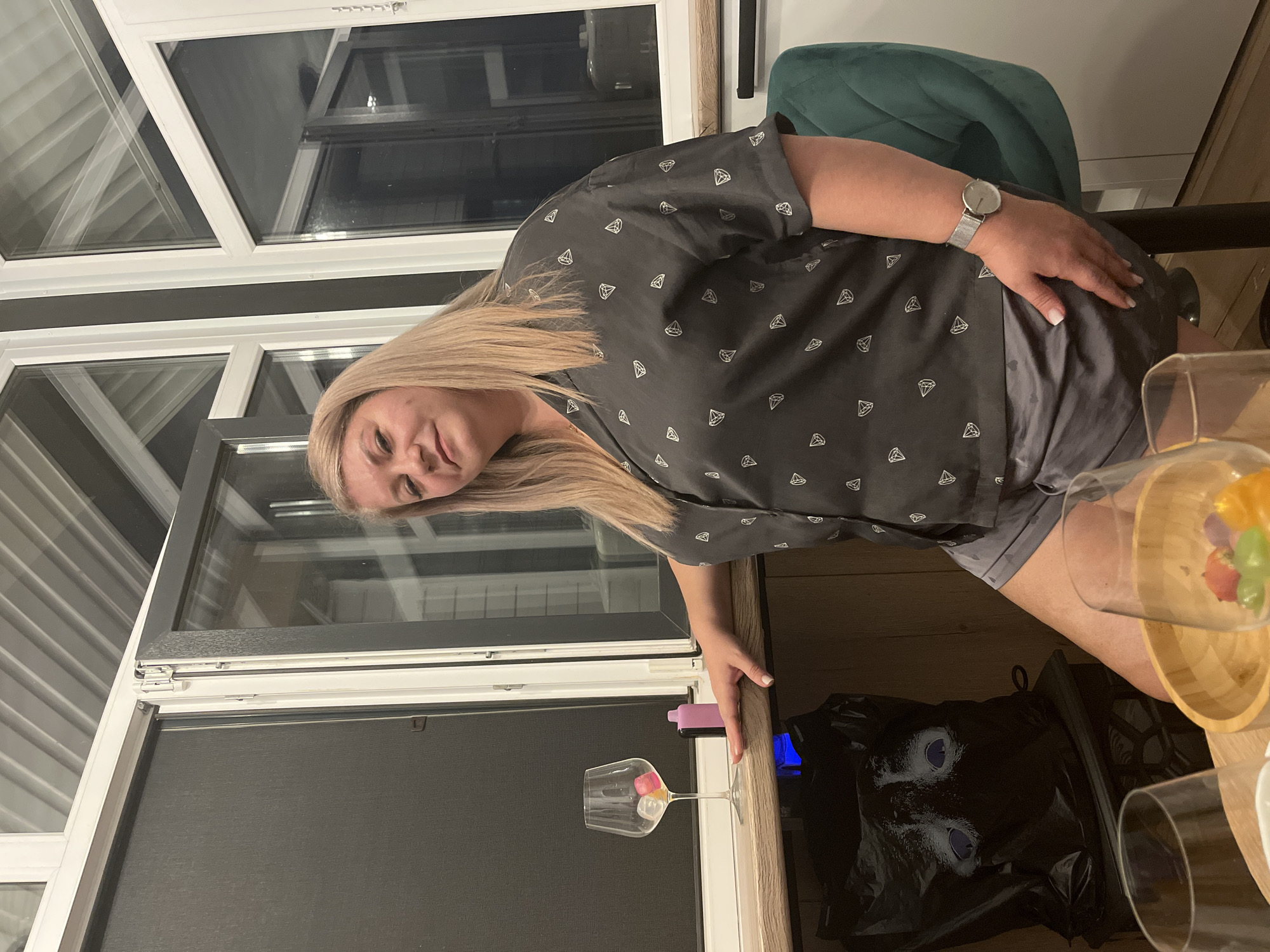 The image depicts an indoor scene in Kamyennoye, Russia, featuring a single adult female in what appears to be a residential or private bar-like setting.
**Subject:**
A female individual, estimated to be between 35-55 years old, with light skin and shoulder-length blonde hair, is seated on a high stool. She is wearing a dark grey short-sleeved collared shirt patterned with numerous small white diamond shapes and dark grey shorts. A silver-colored watch with a round, light-colored face and a mesh band is visible on her left wrist. Her fingernails are manicured with a light polish. Her right hand rests on a wooden counter surface. She is looking directly towards the camera with a neutral expression.
**Environment and Objects:**
The background features multiple white-framed windows. The leftmost window panel has a dark blind or screen drawn. Other window panels show reflections of the interior and an overhead white slatted structure, possibly an awning or ceiling. A dark vertical beam separates some window sections.
On the wooden counter, to the left of the woman's hand, is a clear stemmed wine glass containing several translucent, multicolored cube-shaped objects (likely ice or decorative items) and a small, opaque pink object. Adjacent to the glass is a rectangular, pink and black object resembling a disposable vape pen.
Under the counter, to the left, a black plastic bag with a printed image of stylized animal eyes (blue irises) is visible, resting on a dark stand or small stool. A subtle blue glow emanates from beneath the counter.
In the foreground, on a lower wooden surface, two empty clear glasses and a wooden bowl filled with various colorful fruits (including grapes, berries, and orange segments) are present. A dark green upholstered high-backed stool or chair is partially visible to the right of the woman. The flooring appears dark. The overall lighting is artificial, suggesting evening or nighttime.