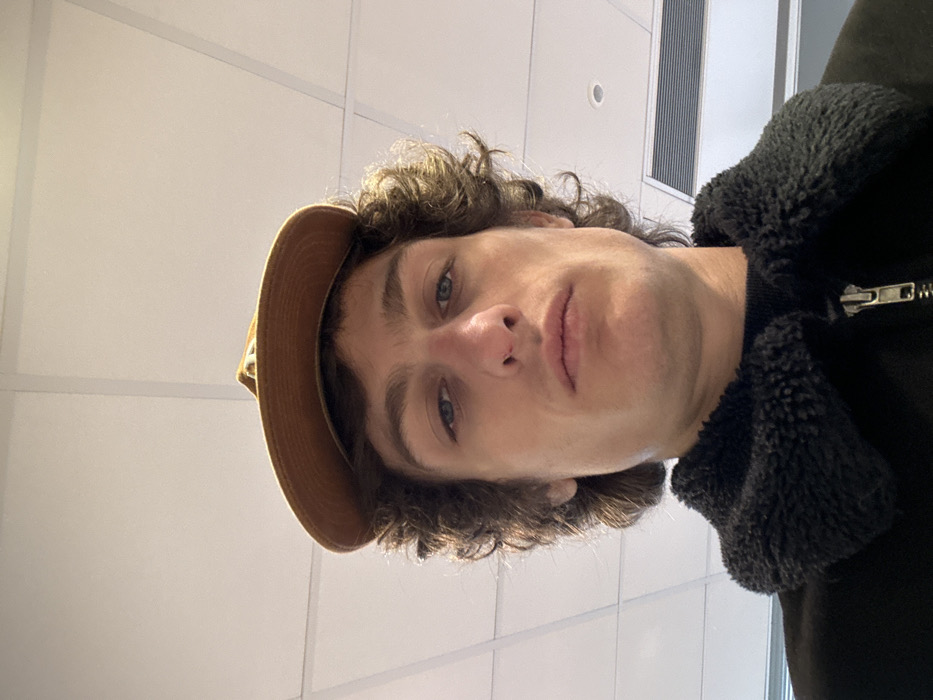 A fair-skinned male individual with curly brown hair and blue eyes is depicted in a close-up, low-angle frontal shot, looking directly at the camera with a neutral expression. He wears a brown baseball cap, positioned backward and slightly elevated, and a dark-colored jacket featuring a prominent, textured black collar with a visible metal zipper pull. The background consists of a white drop ceiling with distinct grid lines, typical of an indoor commercial or public building. A rectangular ventilation grille is partially visible in the upper right quadrant of the image. The specific location within Joinville-le-Pont, France, cannot be determined from the visual evidence presented.