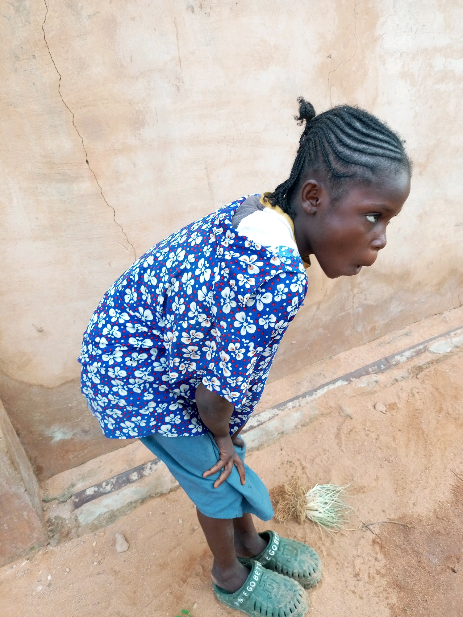 A young Black girl with braided hair and a blue floral shirt leans over, looking down at a small bunch of grass on the dusty ground. She wears blue shorts and green clogs. The background features a cracked, tan-colored wall and a dirt path with some concrete steps. The scene appears to be outdoors in a rural or semi-urban area.