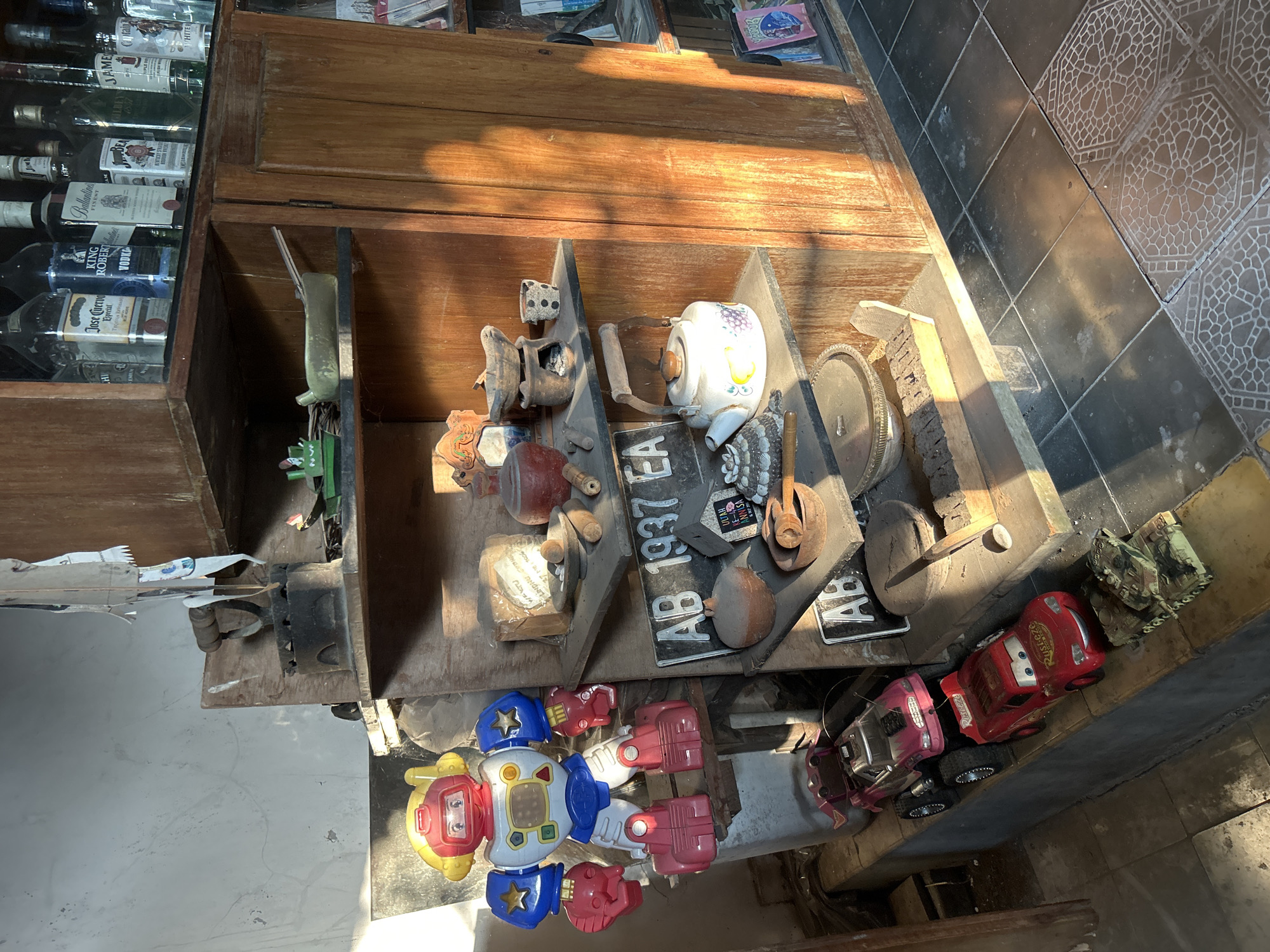 This media file captures a still life scene featuring a collection of diverse objects displayed within a wooden cabinet and on a ledge. The arrangement suggests a curated display of vintage items and toys.
The primary subjects are various objects, including several bottles of alcohol on the left side of the cabinet, brands like Jose Cuervo Especial, King Robert Vodka, and Ballantine's are visible. Inside the cabinet, a variety of items are arranged, such as a white teapot with a grape design, a pair of antique irons, and license plates, one with "AB 1937 EA" clearly legible.
On the ledge below the cabinet, more items are displayed. These include a colorful toy robot, toy cars (one red resembling Lightning McQueen from Cars, and a pink toy car), and a camouflaged toy tank. There are also other assorted trinkets and metal objects.
The scene is set indoors, likely in a shop or a home with a display area. The lighting suggests it's daytime, with sunlight casting strong shadows and highlighting the textures of the wooden cabinet and the tiled floor. The overall mood is one of nostalgic collection and perhaps a touch of playful arrangement due to the toys. The mention of "Banguntapan, Indonesia" provides a geographical context for the setting. There is text visible on the alcohol bottles and the license plates, as well as on the red toy car.
