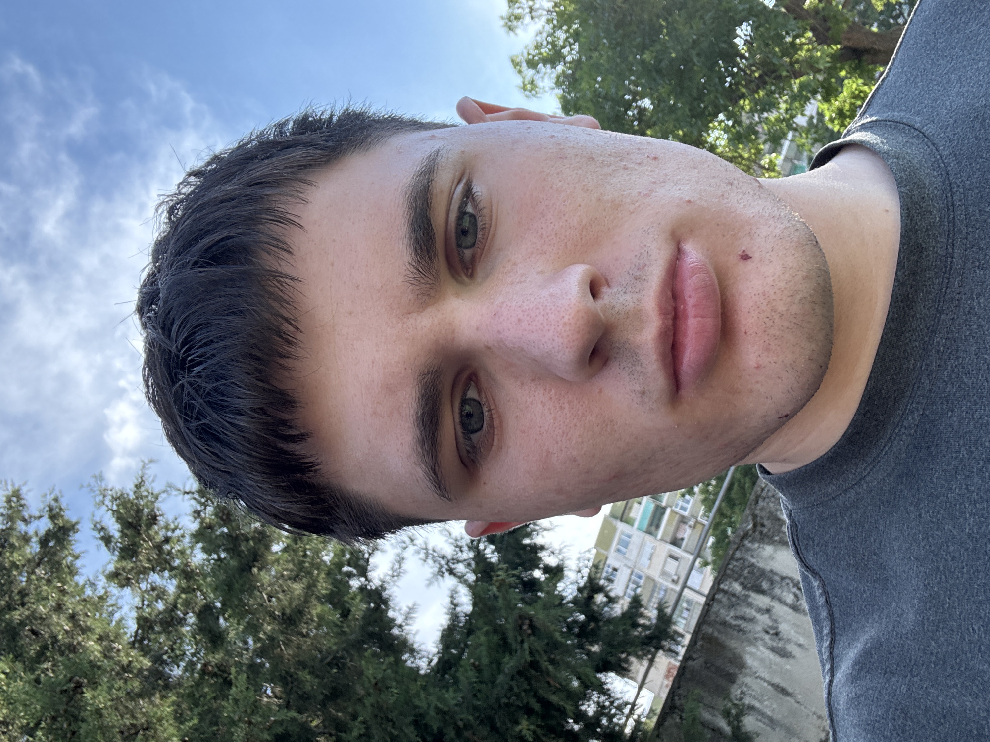 A close-up, waist-up, outdoor selfie of a young adult male with dark, short hair, fair skin with light blemishes, and green eyes. He is wearing a dark gray crewneck t-shirt. The background features a clear blue sky with some white clouds, a large green tree on the left, and a multistory building with light-colored facades and balconies partially visible behind a gray concrete wall on the left. Green foliage from another tree is visible on the right side of the frame. The scene is likely an urban residential area in Stara Zagora, Bulgaria, given the architectural style of the building and the presence of mature trees, indicative of a park or a residential courtyard. The lighting suggests it is daytime, possibly late afternoon, due to the angle of the sun casting light on the subject from the left.