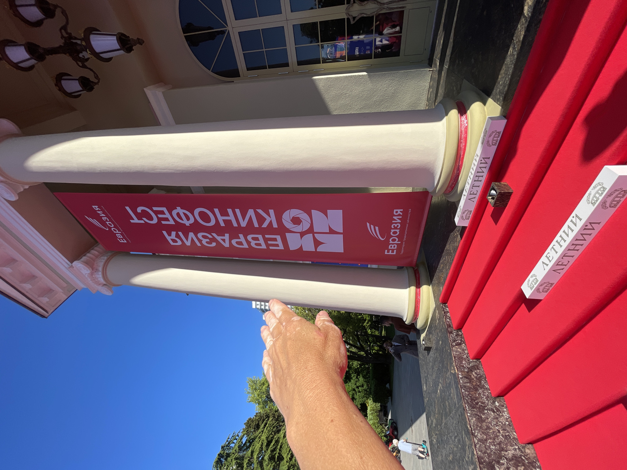 The image depicts the entrance to a building, likely a cinema or cultural center, in Sochi, Russia. A bright red carpet is laid out on steps leading up to the building. Two white boxes with text in Cyrillic script and an illustration of arches are placed on the carpet. A small, dark square object sits on one of the steps. The building features large white columns supporting an ornate pediment, and a red banner with Cyrillic text and a logo hangs between two columns. The sky is clear blue. In the background, trees are visible, and a few people are scattered in the area, including a child on a scooter. A person's hand, with visible vitiligo, is extended towards the banner, suggesting the image is taken from a first-person perspective.