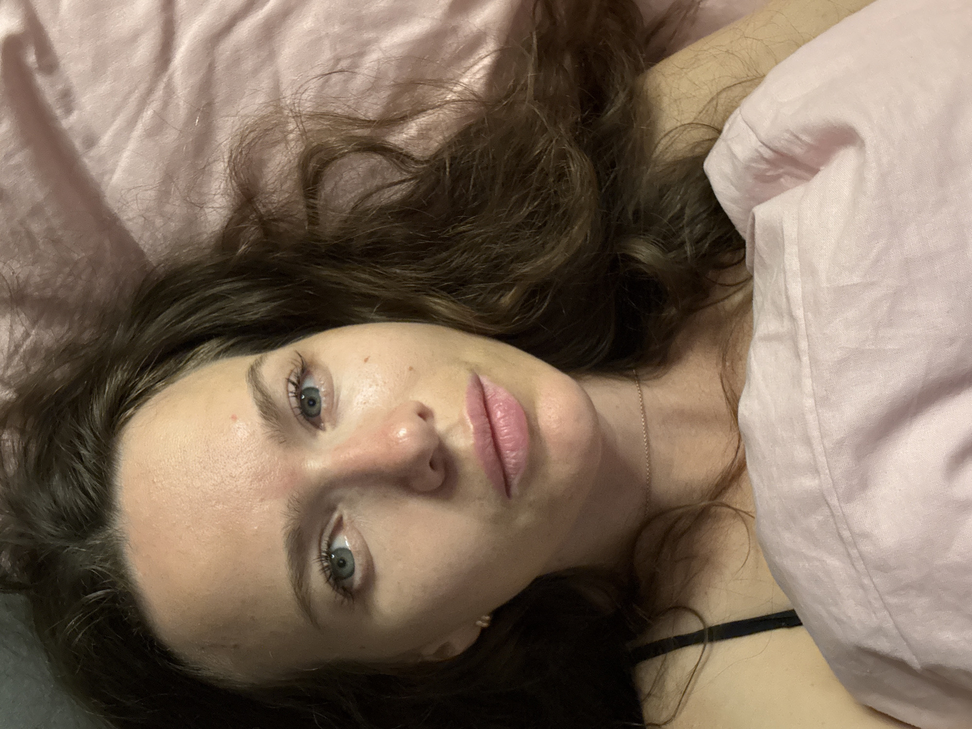 A close-up, overhead shot shows a woman lying in bed, facing the camera. She has long, dark brown wavy hair that is spread out around her. Her eyes are light blue and open, and she is looking directly at the camera. Her lips are slightly parted and appear to be a natural pink color. She is wearing a thin, rose gold necklace around her neck and a black strap is visible on her left shoulder, likely from a tank top or bra. The woman is covered by a pink duvet or blanket. The background consists of the pink bedding, which has a satin-like sheen. The overall impression is of a casual, intimate moment, likely taken in a bedroom. The metadata indicates the image was taken in Khimki, Russia.