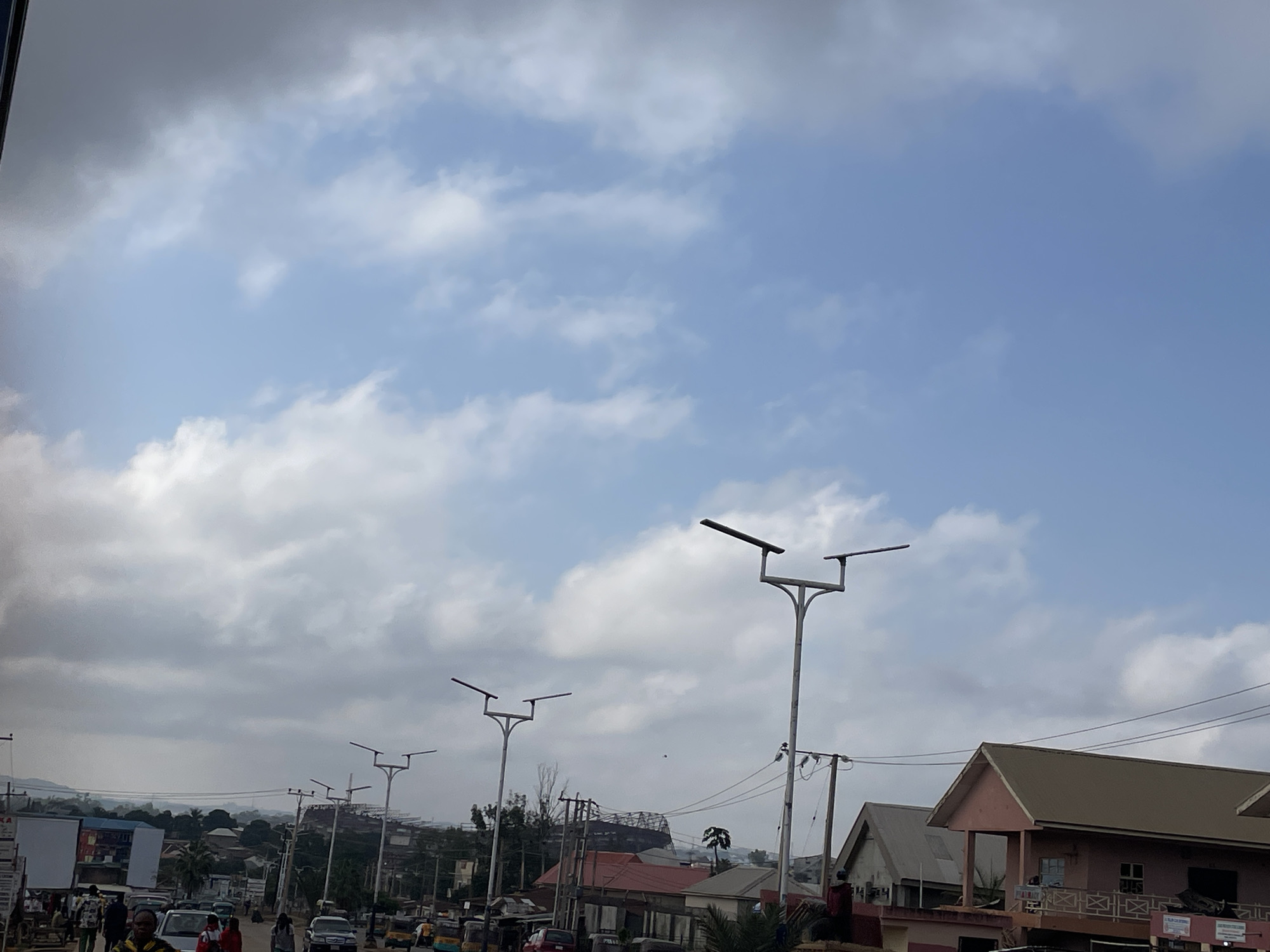 The image captures a street scene in Jos, Nigeria, under a partly cloudy sky. In the foreground, a paved road is visible with several vehicles, including cars and a tricycle, moving or parked. People are walking along the roadside. Overhead, modern streetlights with solar panels line the street. In the background, various buildings are seen, some appearing to be commercial establishments or residential homes, with different architectural styles and roof types. Distant hills are faintly visible through the haze and clouds. The overall impression is of a typical urban environment in Jos.
