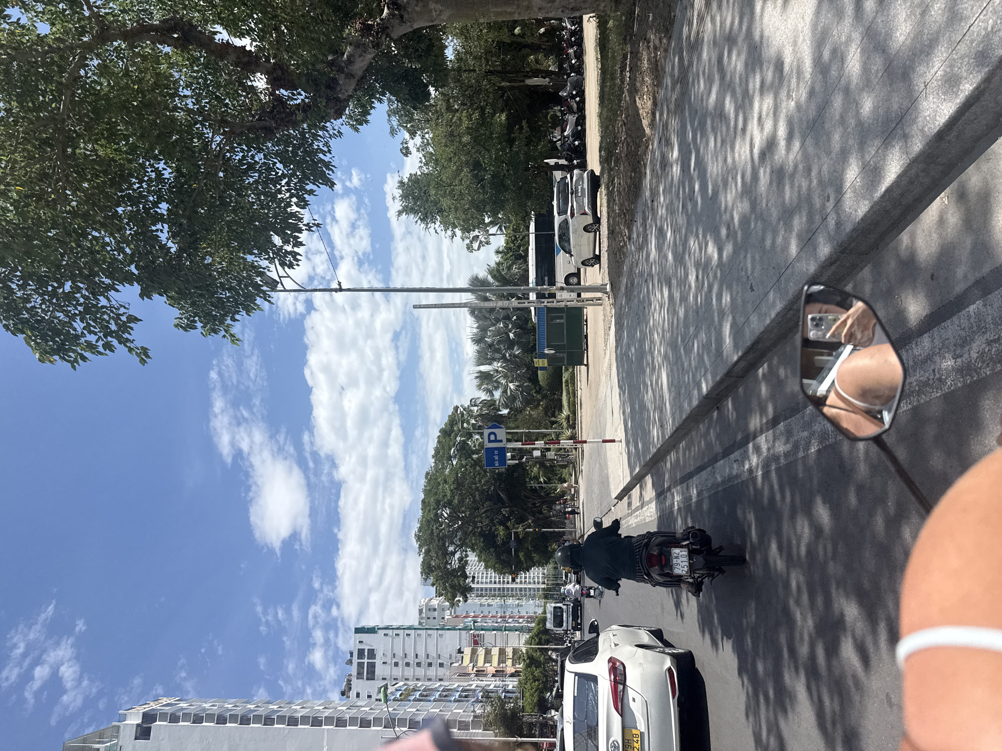 The image depicts a street scene in Nha Trang, Vietnam, on a bright, sunny day with scattered clouds. The perspective appears to be from someone riding a motorcycle, as evidenced by the reflection in the rearview mirror. In the foreground, a person's shoulder and upper torso are visible, wearing a white strap. The rearview mirror shows a reflection of a hand holding a mobile phone, capturing a selfie.
The street itself is paved and lined with a sidewalk. A white car is visible on the left side of the road, with its license plate showing "79H 2.4B". A person wearing a dark green shirt and pants is riding a motorcycle ahead, also with a license plate that reads "79-N2 251.01".
Beyond the street, there are tall, modern buildings, characteristic of a city. To the right, a large tree provides shade over a portion of the sidewalk. Further down the street, traffic lights, street signs, and more trees are visible. There are also parked motorcycles and a white bus in the background. The overall environment suggests a bustling urban area with clear weather.