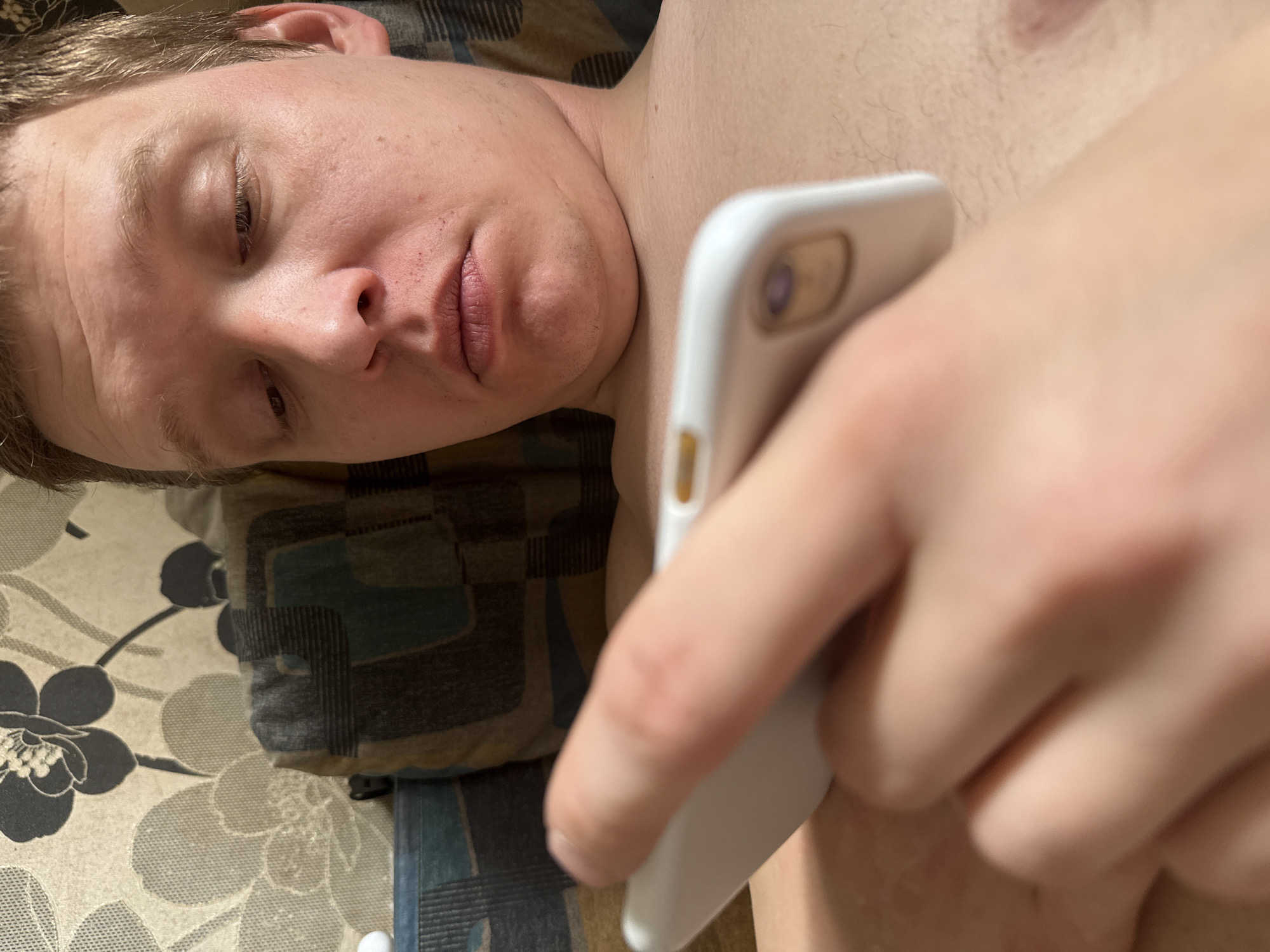 A man is lying down, possibly on a bed. He is holding a white smartphone in his right hand. He appears to be looking at the phone. The background shows a pillow with a floral pattern. The image does not provide information about the location in the city of Dmitrov, Russia.