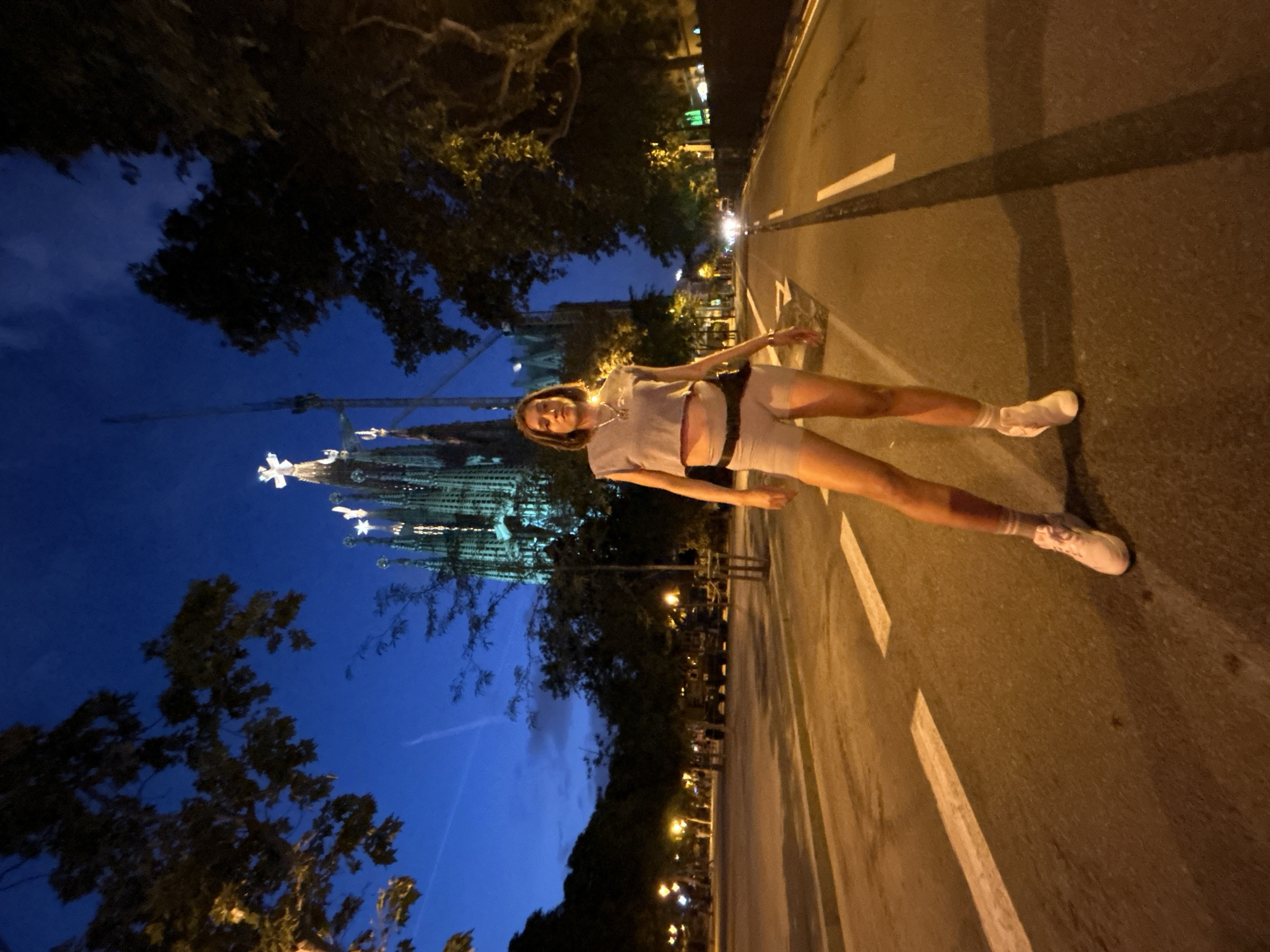 The image depicts a woman standing on a road in Barcelona, Spain, at twilight. She is wearing a grey crop top, light-colored shorts, white socks, and pink sneakers. She has a black fanny pack around her waist. Behind her, the Sagrada Familia, a large basilica, is illuminated against the dark blue sky. Trees line the sides of the road, and streetlights are visible in the background. The road has white lane markings, and there are shadows cast by the trees and the woman. The overall atmosphere is dimly lit, suggesting it is evening or early morning.