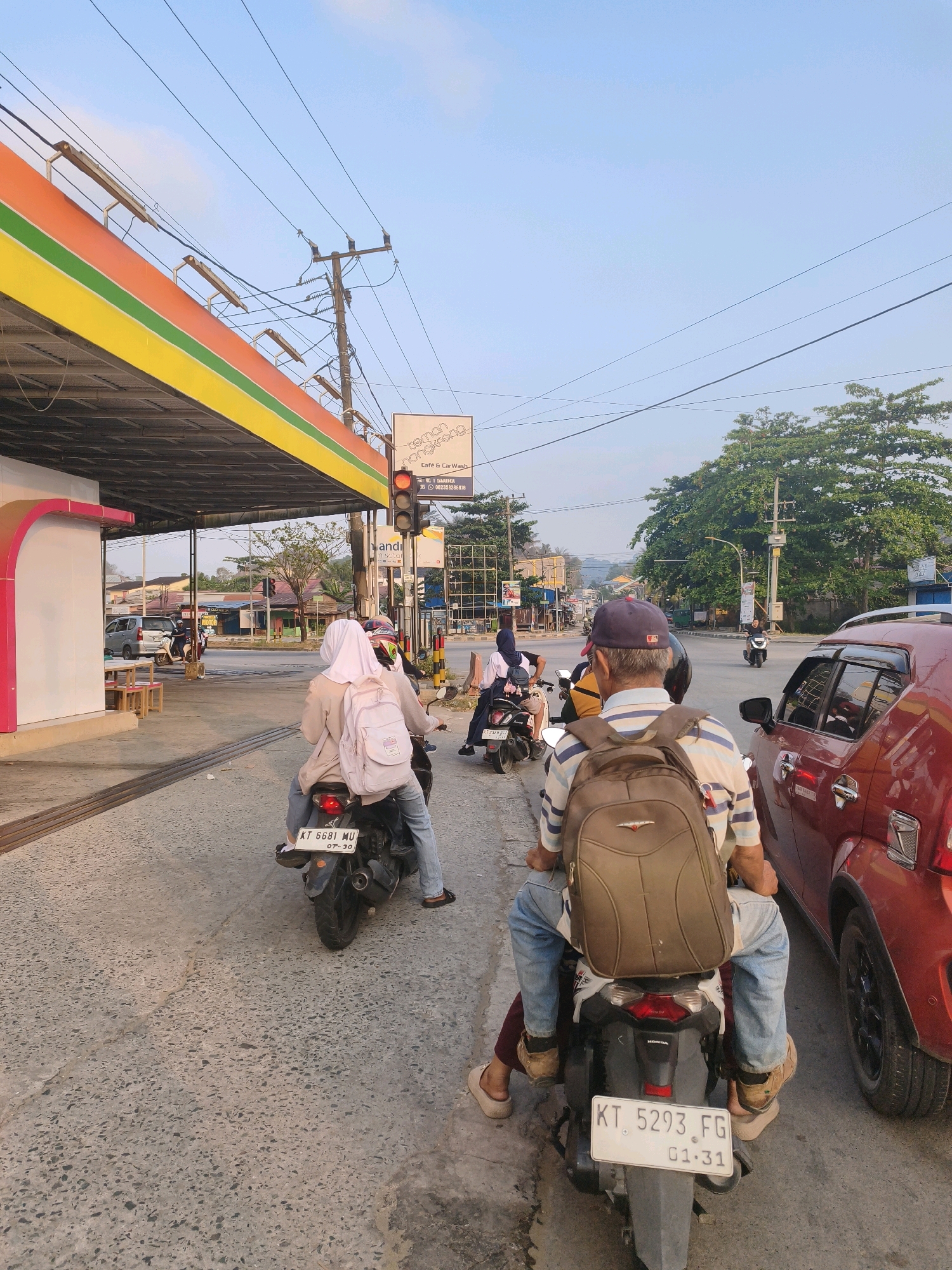 The image depicts a street scene in Samarinda, Indonesia, during daylight hours. In the foreground, a man wearing a blue baseball cap and a striped shirt, carrying a brown backpack, is seated on a motorcycle. He is accompanied by another person behind him, whose legs are visible. To the left of the foreground motorcycle, another motorcycle is seen with two passengers, a woman in a white hijab and a lighter colored jacket, and another person. Both motorcycles are stopped, likely at a traffic light or in traffic.
In the background, several other vehicles, including cars and motorcycles, are visible on the road. A red car is parked to the right of the foreground motorcycle. A gas station canopy, featuring orange, yellow, and green stripes, is present on the left side of the image, with a pink curved structure beneath it. A traffic light is visible hanging above the road, indicating a traffic intersection. Buildings and trees line the street, suggesting an urban or suburban environment. Power lines crisscross the sky. The overall scene captures a moment of daily life in a bustling Indonesian city.