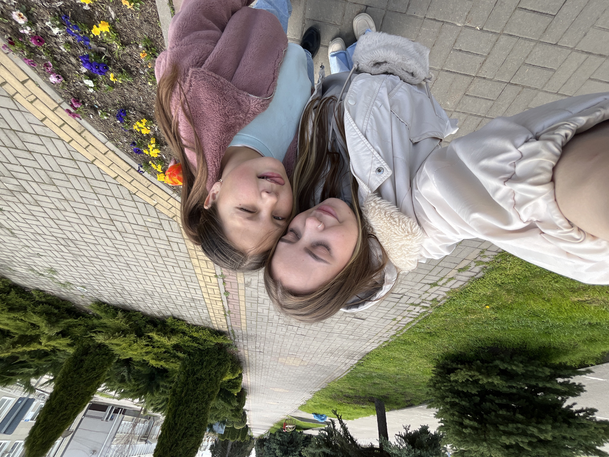 Two young women are posing for a selfie on a brick walkway. One woman wears a pink jacket and a light blue shirt, while the other wears a light-colored jacket with a fur-lined hood. They are standing next to a flower bed with colorful flowers on the left and a green grassy area with trees on the right. The setting appears to be a street in Novopokrovskaya, Russia. A building is visible in the background.
