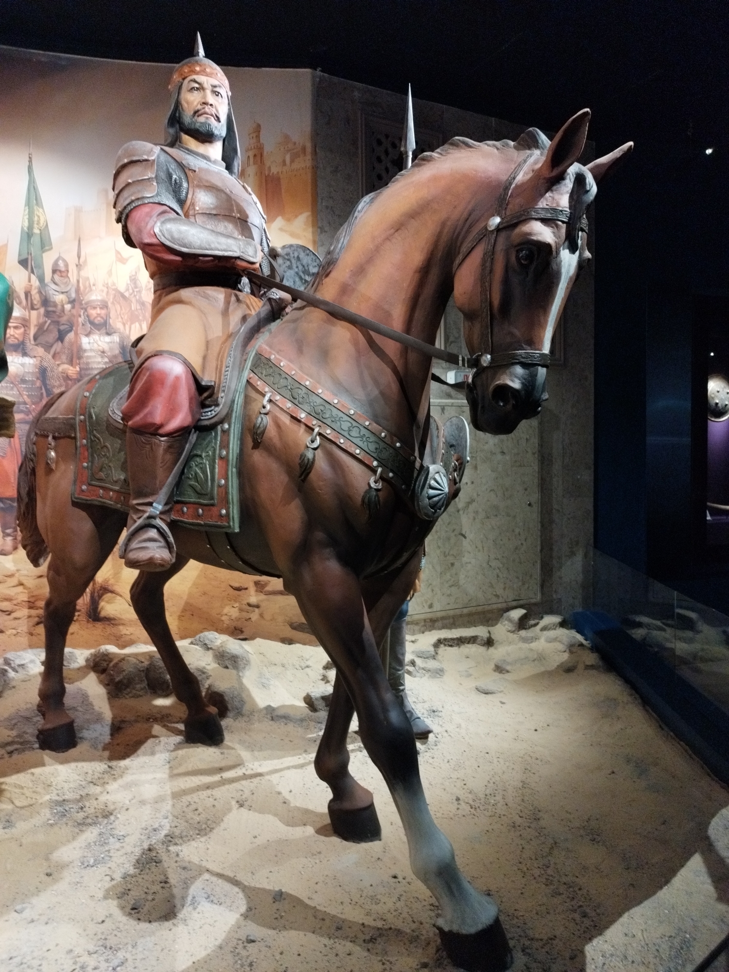 A life-sized statue of a man on horseback is displayed in a museum exhibit. The man is wearing historical armor and a helmet. The horse is brown and adorned with decorative tack. The backdrop depicts a scene of soldiers and a city, suggesting a historical battle or procession. The exhibit appears to be staged on a sandy floor with rocks. The lighting is dim, with spotlights illuminating the central figures. The scene is viewed from a low angle, emphasizing the imposing presence of the statue. The overall impression is of a well-preserved historical display.