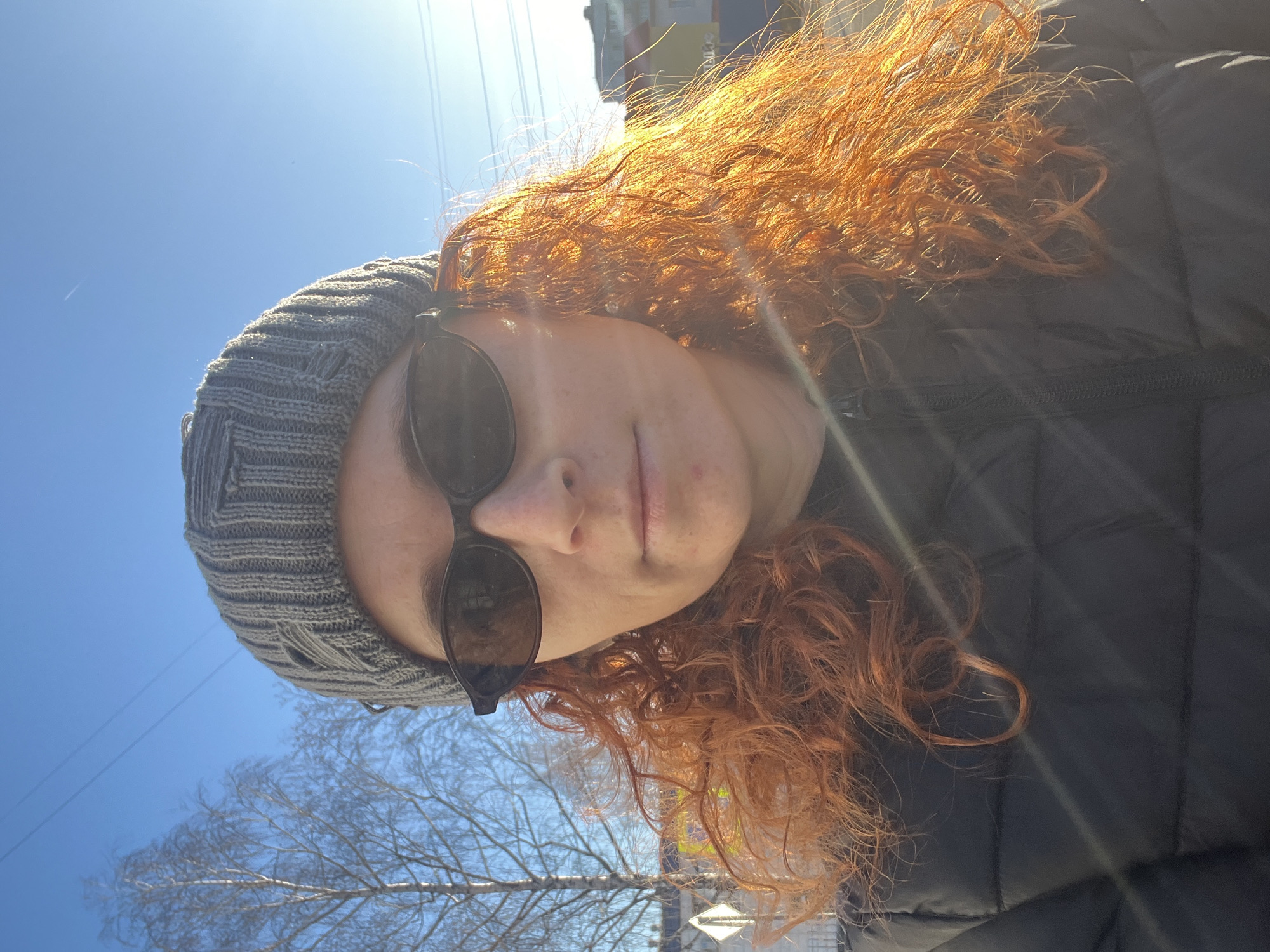 A rotated outdoor portrait shows a person with long, voluminous, curly reddish-orange hair, wearing a grey knitted beanie and dark rounded sunglasses. The person is looking directly at the camera, with a neutral to slightly smiling expression. A black puffer jacket covers the subject's torso.
The setting is outdoors in Chaikovsky, Russia, under a clear, bright blue sky. Strong sunlight creates a prominent lens flare across the image and intensely illuminates the subject's hair. In the background, faint power lines stretch across the sky, and bare tree branches are visible, suggesting late winter or early spring. Distant buildings can be vaguely discerned on the horizon. The bright light indicates it is daytime, likely midday or afternoon. No discernible text is visible in the image.