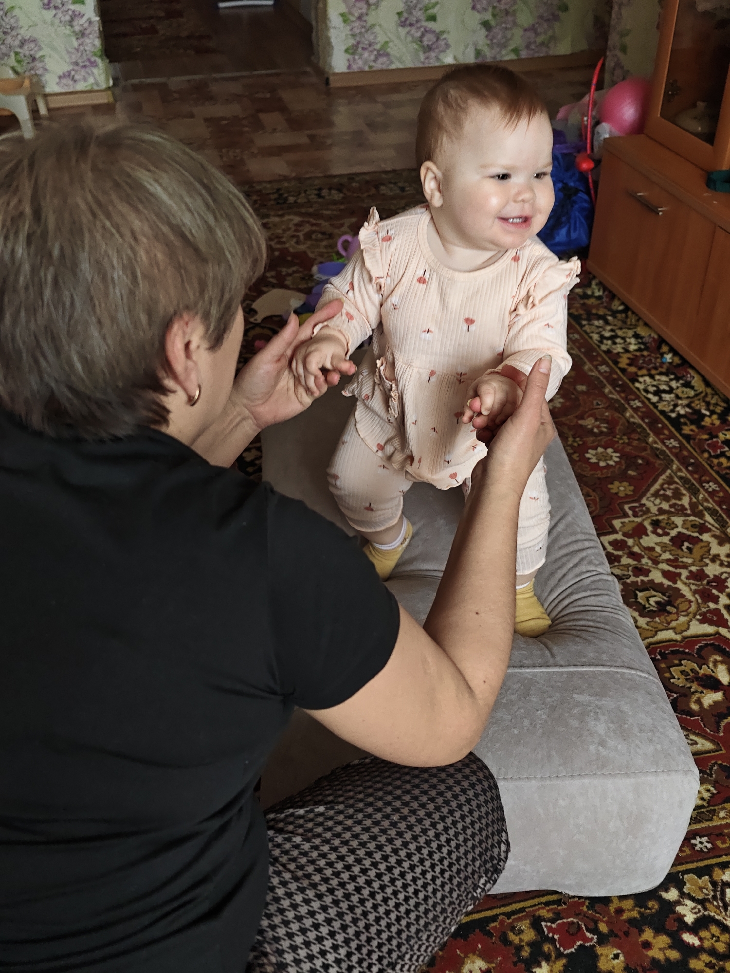 The photograph was taken in a room in Уральское, Russia. An elderly woman with short, gray-streaked hair and gold hoop earrings, wearing a black t-shirt and houndstooth-patterned pants, is seated. She is holding the hands of a smiling baby wearing a pink outfit with a ruffled collar and yellow socks. The baby is standing on a gray, rectangular ottoman placed on a patterned rug. In the background, there's a wooden cabinet, a pink balloon, and a wall with floral wallpaper. The room appears to be a living space within a residential building.