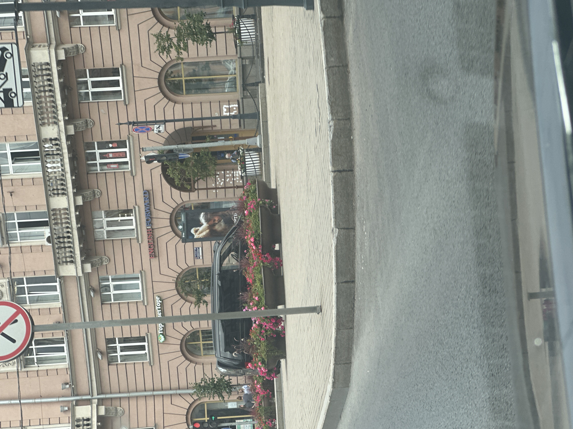 A black van is parked on a city street in Saint Petersburg, Russia, with a row of potted plants in front of it. The van is facing to the left, and its windows are tinted. The potted plants are filled with pink and red flowers. In the background, there is a building with an arched entryway. A traffic light is visible on the left side of the image.