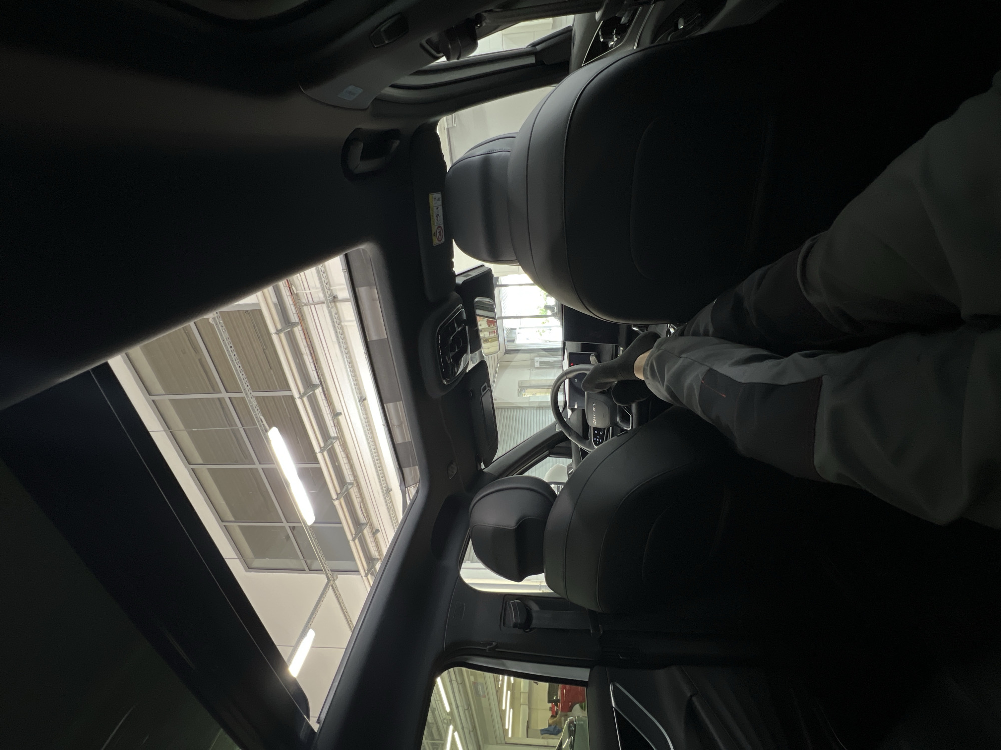 The image captures the interior of a modern vehicle from the perspective of the rear passenger seat, looking forward. A person's legs, clad in grey and black athletic pants and black socks, are extended and resting on the dashboard or front console. The vehicle features a black interior, including black leather front seats, a multi-function steering wheel partially visible, and a panoramic sunroof, which is open. An airbag warning label is affixed to the passenger-side sun visor.
Through the open sunroof and vehicle windows, the interior of a brightly lit, structured building is visible. The building's ceiling incorporates large rectangular windows, and below these, multiple rows of horizontal metal tracks support linear fluorescent light fixtures. In the background, to the left, a portion of a reddish vehicle is discernible. To the right, a large roll-up door or gate is visible. The environment suggests an indoor commercial or industrial space, likely a car dealership showroom or service center, located in Izhevsk, Russia.