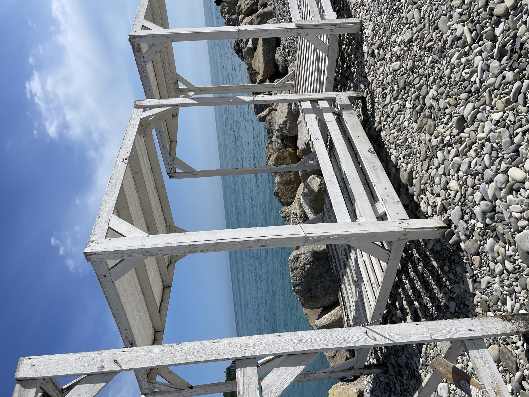 The image captures a scene on a pebble beach in Olginka, Russia. The foreground features weathered white wooden structures, possibly remains of beach infrastructure like sunshades or changing booths. The structures cast shadows onto the grey pebbles covering the beach. Beyond these, large rocks separate the beach from the calm, light blue sea. The sky is a clear blue with some scattered clouds visible above the sea. The entire image is oriented in a portrait-style view.