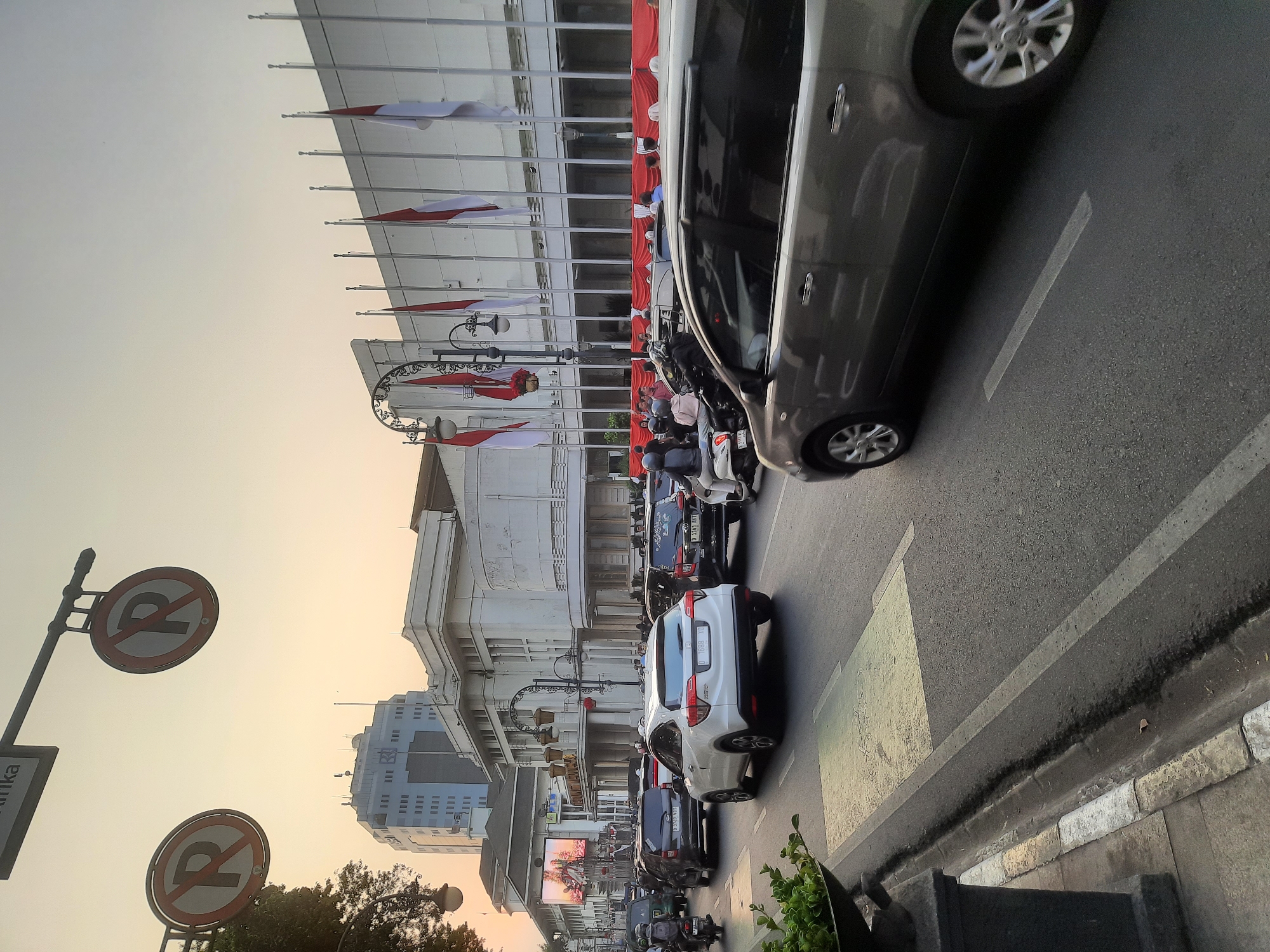 This is a medium-shot, eye-level, outdoor, vertical, natural-light photograph taken in Bandung City, Indonesia. The image depicts a street scene with moderate traffic congestion, likely during the late afternoon or early evening as indicated by the warm, soft lighting.
In the foreground, the right side of a gray SUV is visible, with its rear wheel and door handle in focus. The SUV is in motion, moving towards the right of the frame. To the left of the SUV, the road surface is visible, with a pedestrian crossing marked by yellow and white lines. A portion of a dark gray planter with green foliage is also present on the far left, partially obscuring the sidewalk.
The middle ground is dominated by a line of vehicles, including cars and motorcycles, navigating the street. A white Honda SUV is prominently positioned in the center, its rear facing the viewer. Behind it, other cars are stopped or moving slowly. Several individuals are visible on motorcycles and as pedestrians.
The background features several buildings with architectural styles indicative of a historical city center. A tall, modern blue-tinted building stands in the distance on the left. In the center, a long, white, colonial-era building with columns and large windows is visible, adorned with numerous national flags (red and white) on flagpoles along its facade. A large digital screen displaying an advertisement is mounted on the side of a building on the left. Overhead, two "No Parking" signs with a red circle and a diagonal line over a "P" symbol are suspended by a metal pole. The sky is a pale orange-yellow, suggesting either sunrise or sunset. The overall impression is a bustling urban environment with a blend of historical and modern architecture under a warm sky.