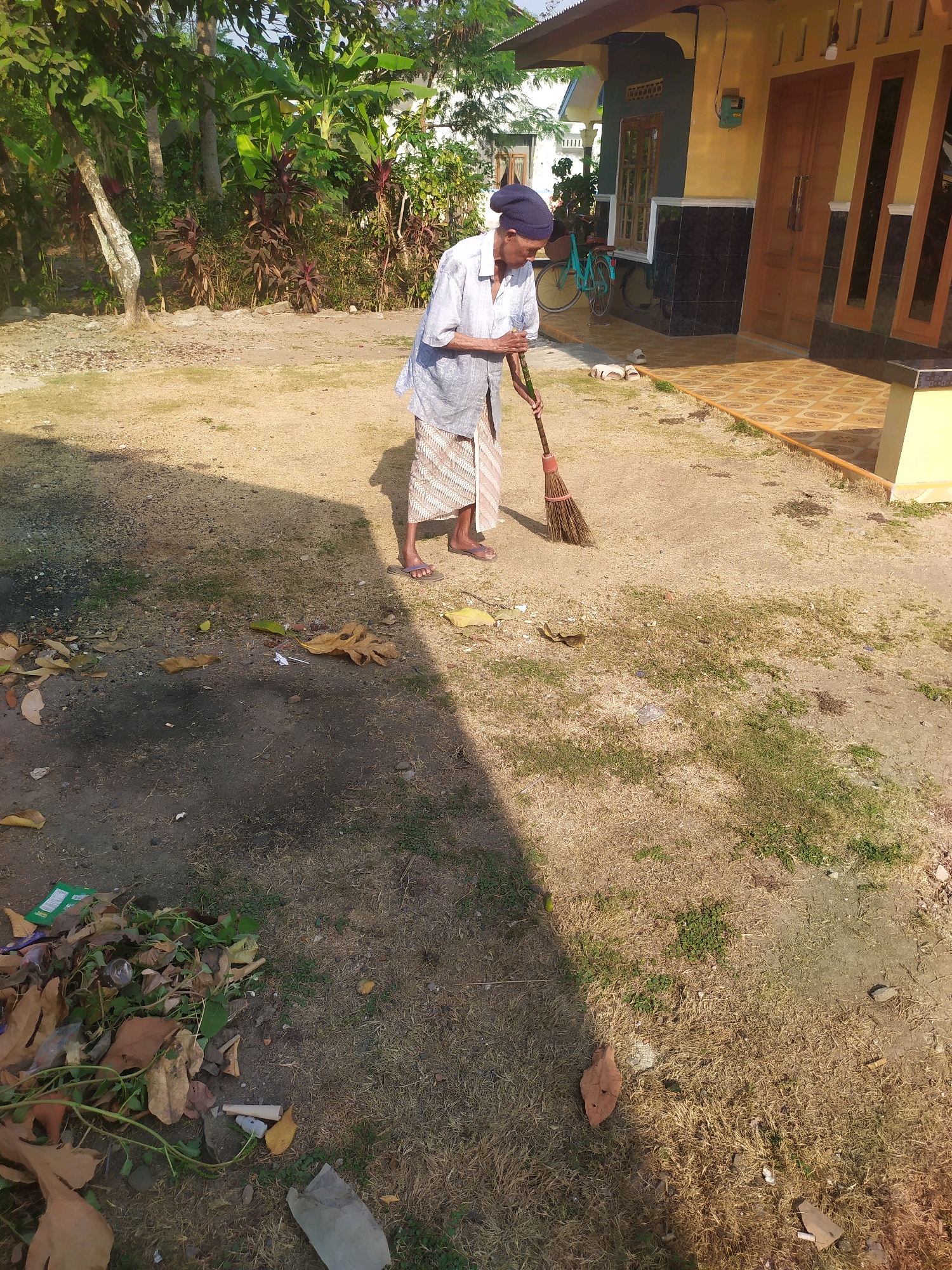In the village of Desa Penggalang, Kecamatan Adipala, Indonesia, a person wearing a blue headscarf, a light blue long-sleeved shirt, and a patterned sarong is sweeping the sandy ground with a broom. The person is positioned in front of a yellow and dark tiled house. Behind the house, there is lush green foliage and trees. A teal bicycle is leaning against the house. The ground is covered in dry grass, dirt, and fallen leaves. In the foreground, to the left, there is a pile of debris including dried leaves, green vines, and some plastic waste.