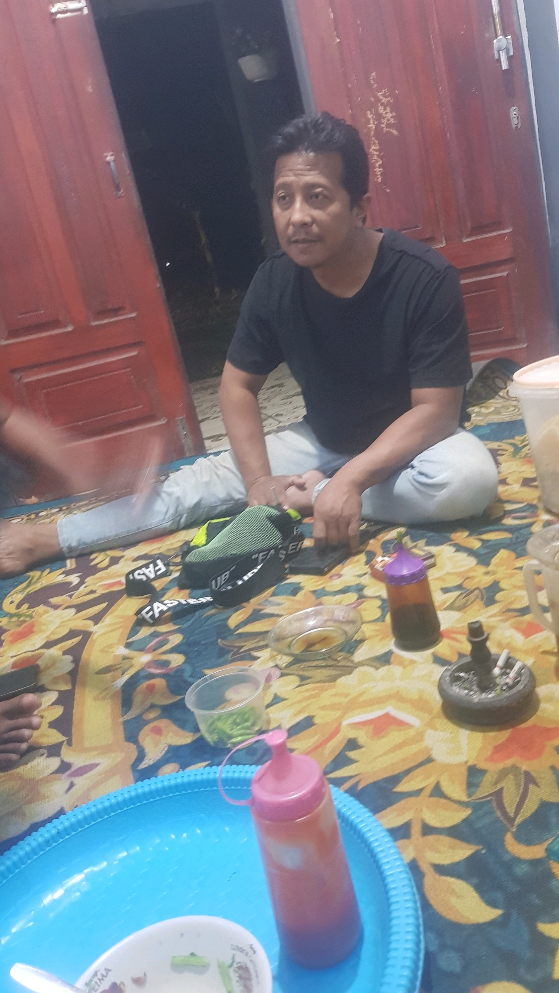 In Bakalan, Indonesia, a man in a black t-shirt and light-colored pants sits on a colorful patterned rug. He is looking to his left, with his left hand resting on his lap. Next to him are several objects: a green and black shoe, a black smartphone, a small glass bowl, a bottle of sauce, a small plastic container with green chili peppers, and an ashtray with cigarette butts. A large blue plastic tray with a white bowl and a spoon is in the foreground. Behind the man are red doors, partially open, revealing a dark interior. The lighting suggests it is nighttime.