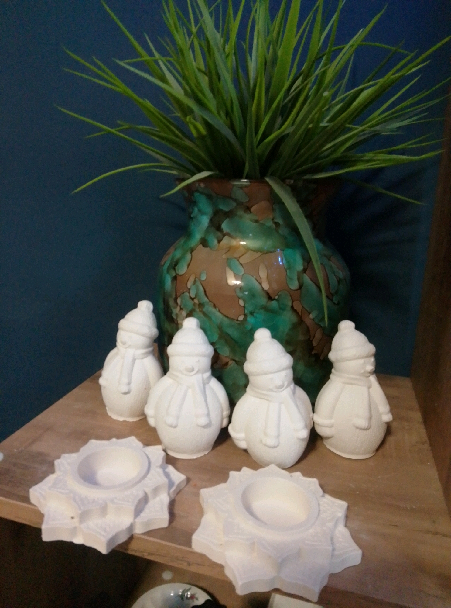 A medium shot captures a still life arrangement on a wooden shelf against a dark blue wall. In the background, a large, ornate vase with a green and brown glaze holds a vibrant green artificial plant. In front of the vase, four unpainted ceramic snowman figures stand in a row. To the left and right of the snowmen, two unpainted ceramic candle holders with star-like designs are placed on the shelf. The lighting is warm and appears to be coming from the right, casting soft shadows. The image does not provide any information or context that would allow for determining its location in the city of Wedel, Germany.
