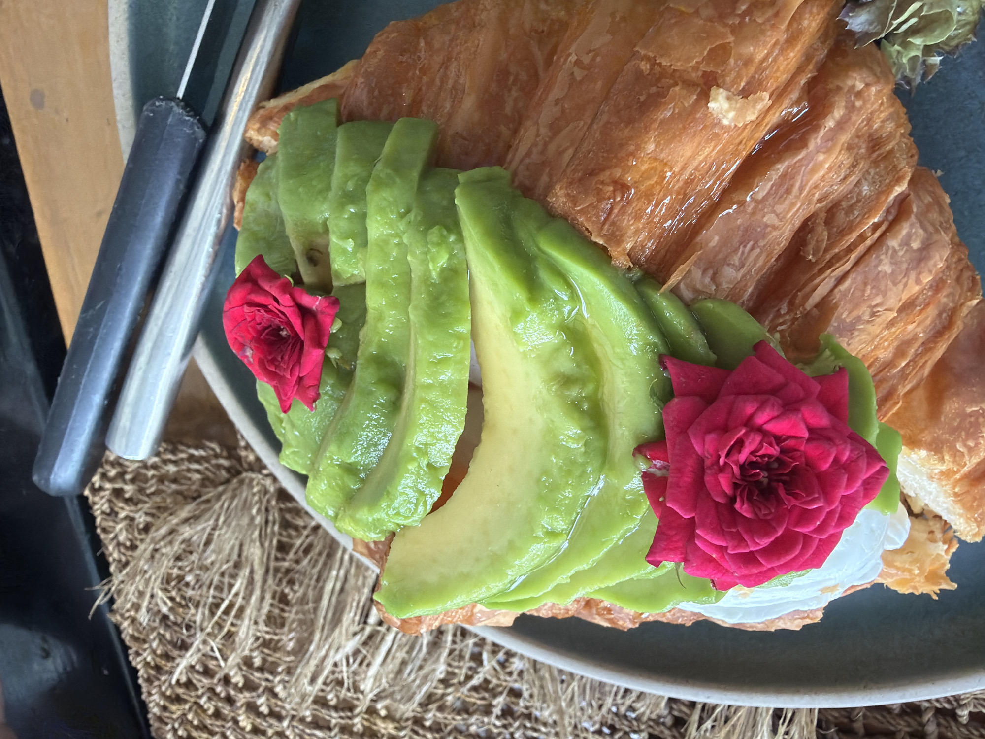 A close-up, overhead shot depicts a gourmet croissant topped with sliced avocado and two vibrant red roses, presented on a grey plate. The croissant, golden brown and flaky, is partially visible. The avocado slices are arranged in a fanned pattern, with a dollop of white cream beneath them. The roses, intricately petaled, add a splash of color to the dish. In the background, a rough, woven placemat is visible, alongside what appears to be cutlery. The scene is well-lit, highlighting the textures and colors of the food. The context suggests a leisurely meal, possibly at a cafe or restaurant in Baan Bo Phut, Thailand.