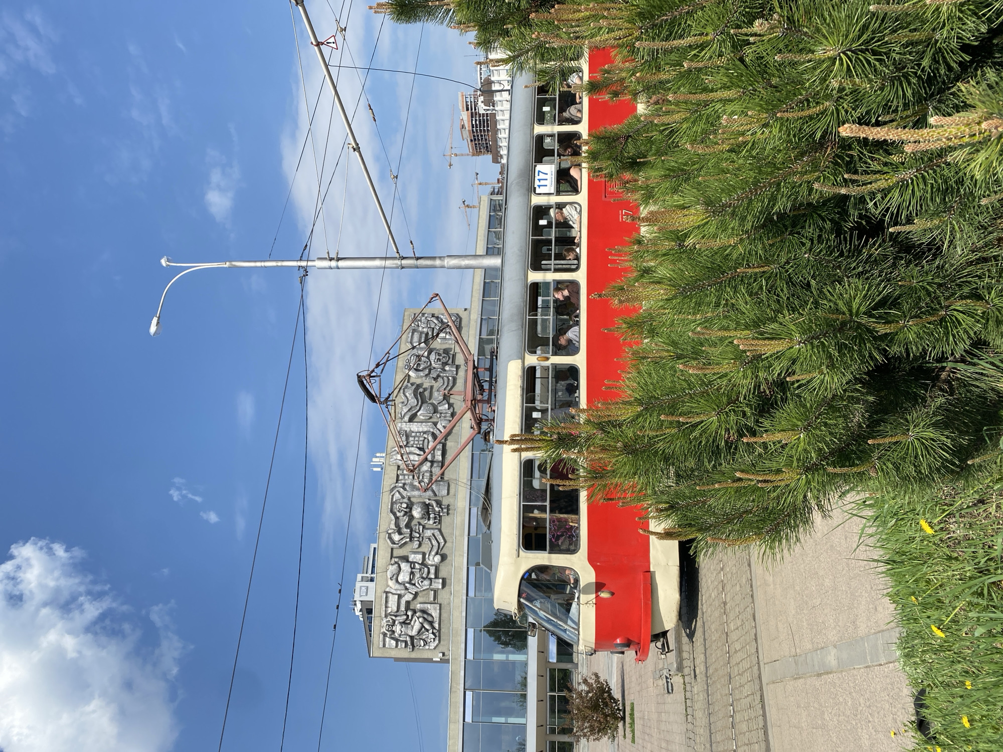 The image captures a street scene in Yekaterinburg, Russia, featuring a red and beige tram filled with passengers. The tram is situated next to a building adorned with a large, concrete Soviet-era relief sculpture. Overhead, wires connect the tram to the power grid. A street lamp stands tall against a blue sky with scattered clouds. A conifer tree partially obscures the tram, adding greenery to the urban environment. In the distance, construction cranes indicate ongoing development in the city. The ground is paved, suggesting a sidewalk or pedestrian area.
