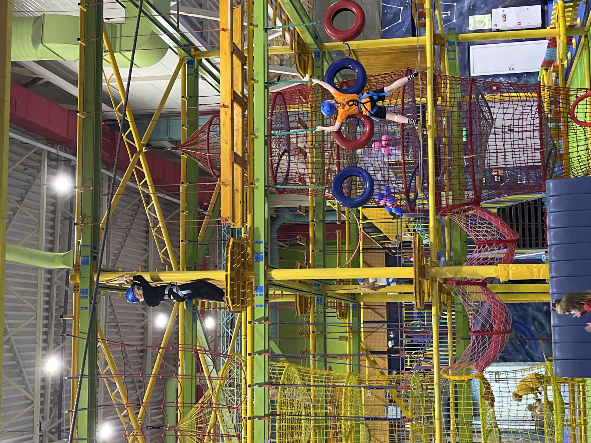 The image depicts an indoor playground, likely located in the city of Krasnoyarsk, Russia. The playground features an intricate network of interconnected structures, including rope bridges, climbing nets, tire obstacles, and platforms. Two individuals are actively engaged in the play activities. One person, wearing a blue helmet and dark clothing, is positioned on a rope bridge. Another person, wearing an orange shirt and a blue helmet, is suspended between tires. The environment is brightly lit with visible ductwork and a high ceiling. The playground's structures are predominantly constructed with yellow and green supports. The foreground includes a glimpse of another person standing near blue padded structures.