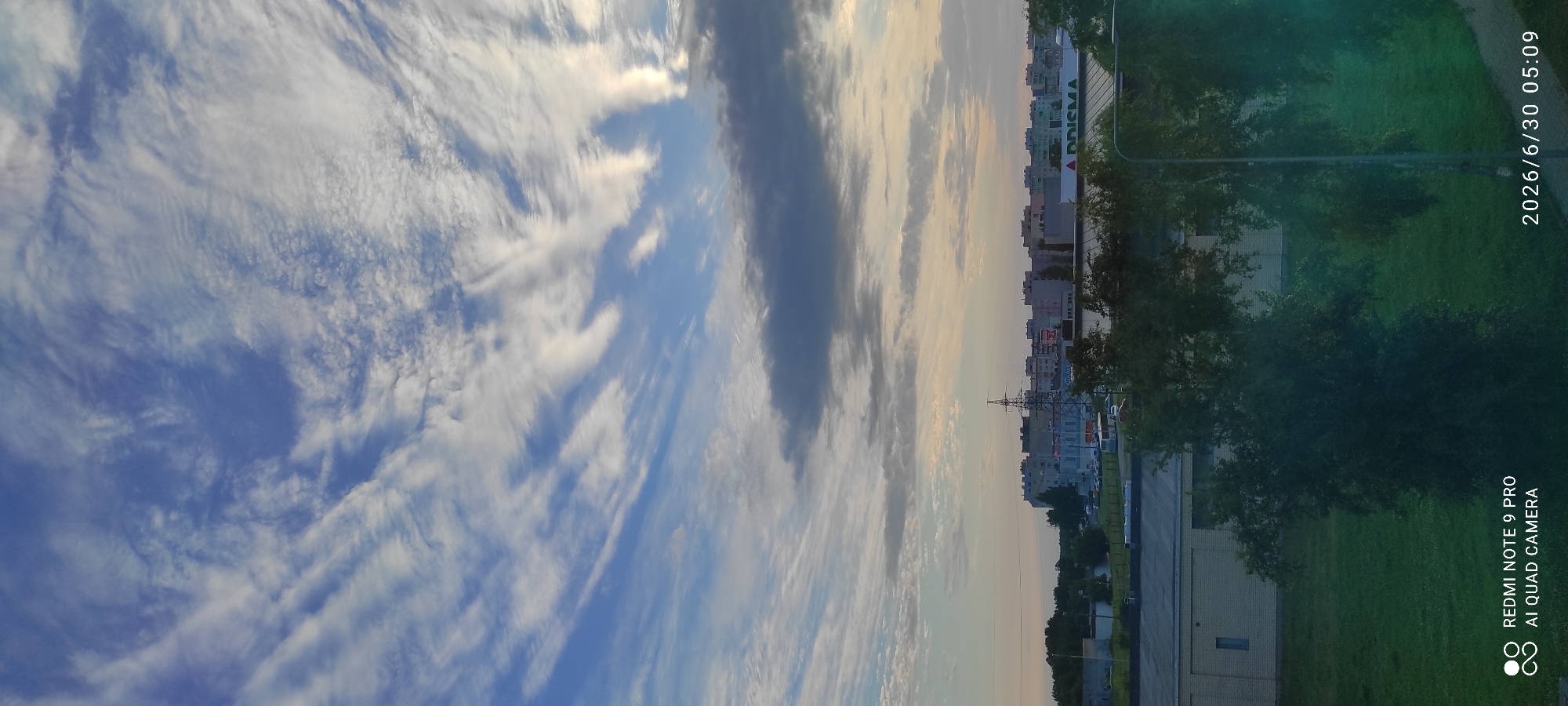 The image shows a wide shot of a cityscape under a dramatic sky. The sky is filled with wispy, elongated white clouds against a blue background, with darker, thicker clouds forming a band across the middle distance. In the lower half of the image, the scene transitions to a view of buildings, trees, and a grassy area. A row of apartment buildings with varied colored facades stretches across the horizon. In front of the buildings, a large retail store with a green roof and a visible "PRISMA" sign is prominent. There are also some smaller industrial-looking buildings, a tall power line pylon, and parked cars. Green trees and foliage occupy the foreground and obscure some of the lower buildings. A street lamp is visible on the right side of the image. The bottom left corner displays a "REDMI NOTE 9 PRO AI QUAD CAMERA" watermark, and the bottom right corner shows a date and time stamp: "2026/6/30 05:09". The overall impression is of an urban environment during daytime, possibly early morning or late afternoon due to the lighting and sky conditions.