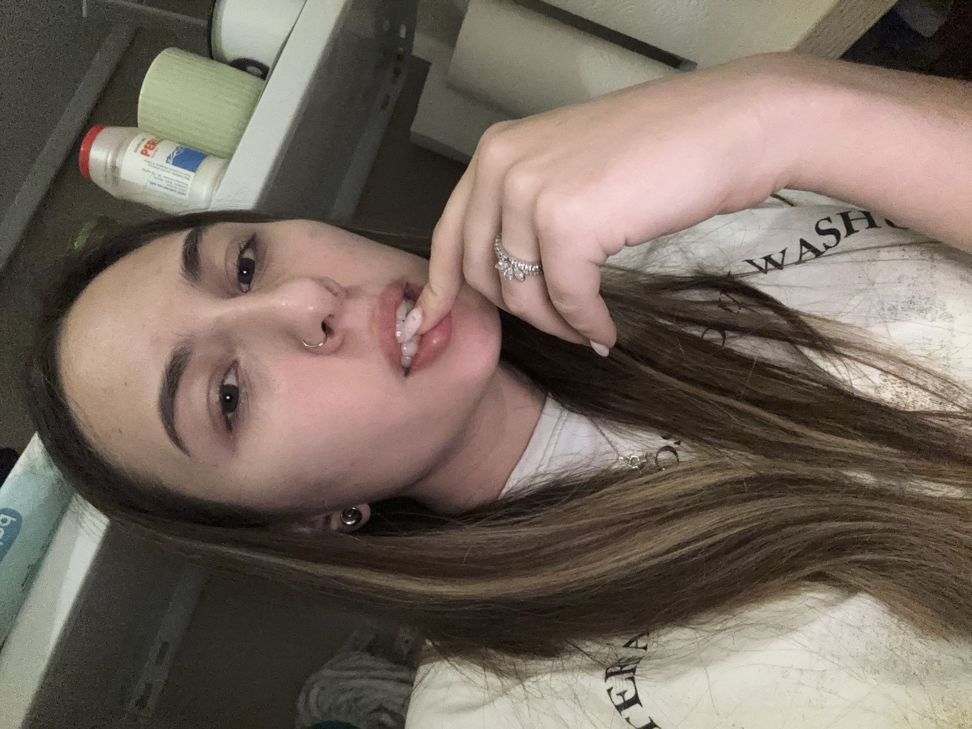 A young woman is positioned at an unusual angle, likely due to the photograph being taken with a phone camera. Her long, highlighted hair is partially obscuring her shirt, which features a printed design. She has a nose piercing and is wearing earrings. Her left hand is near her mouth. A ring is visible on her finger. The background is slightly blurred, showing a shelf with objects like a detergent bottle and containers. The location is not determinable from the image.