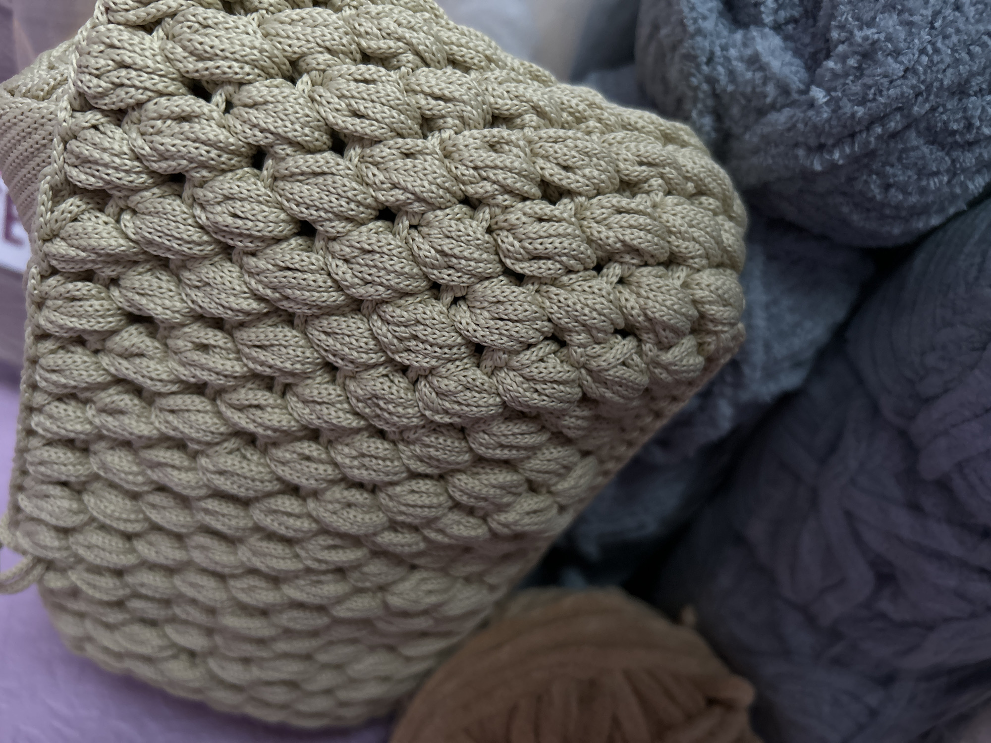 The image presents a close-up view of several balls of yarn in varying textures and colors. In the foreground is a beige yarn ball that has been crocheted with a textured, almost bubble-like pattern. To the right, a ball of gray yarn with a fluffy, plush texture is visible. Below the beige yarn is a small portion of a brown yarn ball. The yarns are resting on what appears to be a lavender-colored surface. There is no indication of Saint Petersburg or any identifiable city location in the image. There are no people, actions, or interactions visible. The scene is limited to the display of the yarn balls on a colored surface.