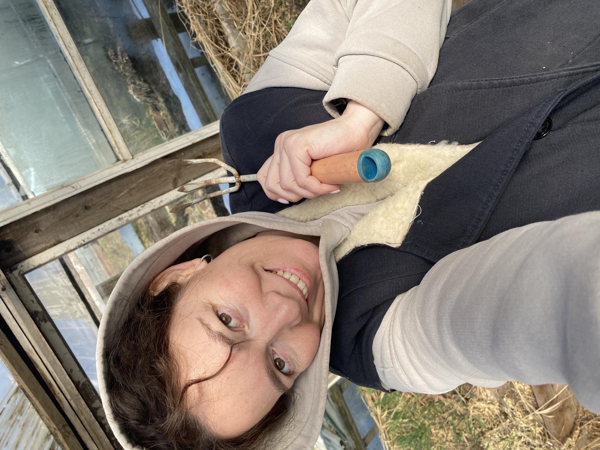 A woman is kneeling inside a weathered greenhouse. She has brown hair and green eyes, and is wearing a hooded jacket with light grey sleeves and a dark blue body lined with light-colored fleece. She is holding a small, three-pronged gardening tool with a wooden handle that is blue at the end. The greenhouse has a wooden frame and glass panes, with a view to the outside showing grass and possibly other plants. The location is likely in or around the city of Ольховка, Russia.