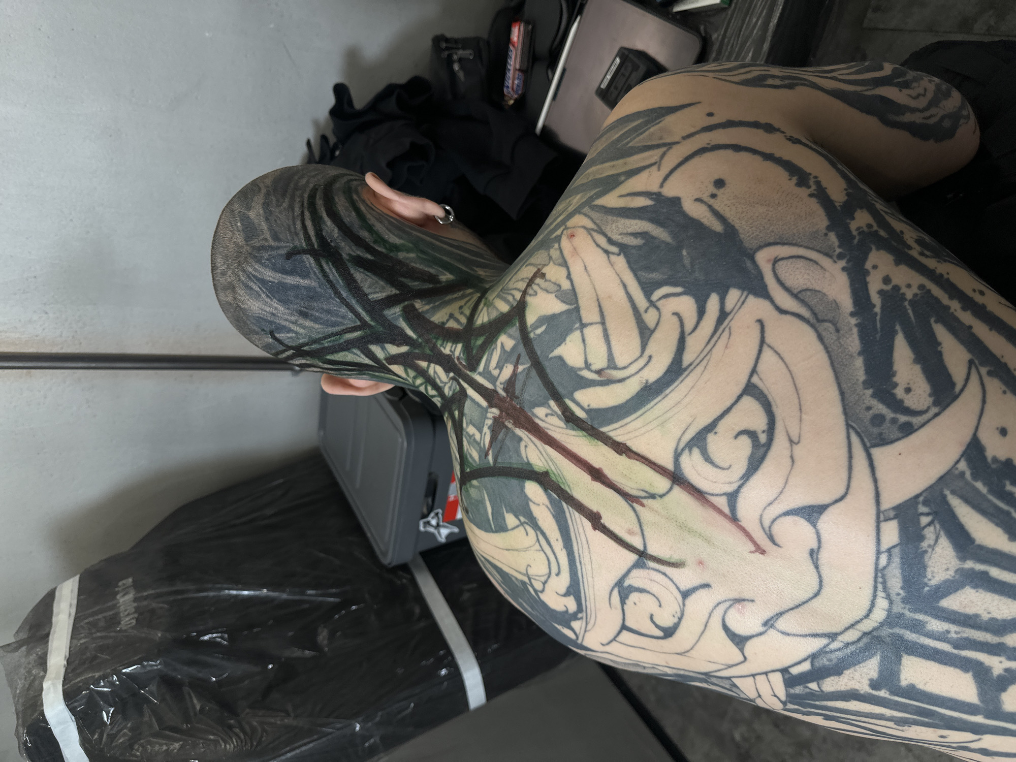 The image shows a person from a slightly overhead, back, and side perspective, likely within a tattoo studio in Moscow, Russia. The individual displays extensive, intricate tattoos in a Japanese-inspired style, covering their shaved head, neck, back, and left arm. The head features dark, organic patterns, possibly representing smoke or waves, while the back and arm showcase a complex design including the outline of a demon or oni mask with horns, surrounded by shaded black and grey patterns with some dark green/blue accents. Red lines are distinctly visible on the neck and upper back, suggesting either fresh red ink or irritation from a recent tattooing session. A small silver hoop earring is present in the person's left ear.
In the background, dark clothing items are visible, alongside a grey container, possibly a tattoo power supply or supply case. The floor and a surface on the left are covered with black plastic sheeting, and the word "dystopia" is visible on a white tape securing the plastic. The setting, with its specific equipment and ongoing body art, strongly indicates an active tattoo session or one that has recently concluded. The lighting is artificial, consistent with an indoor working environment.