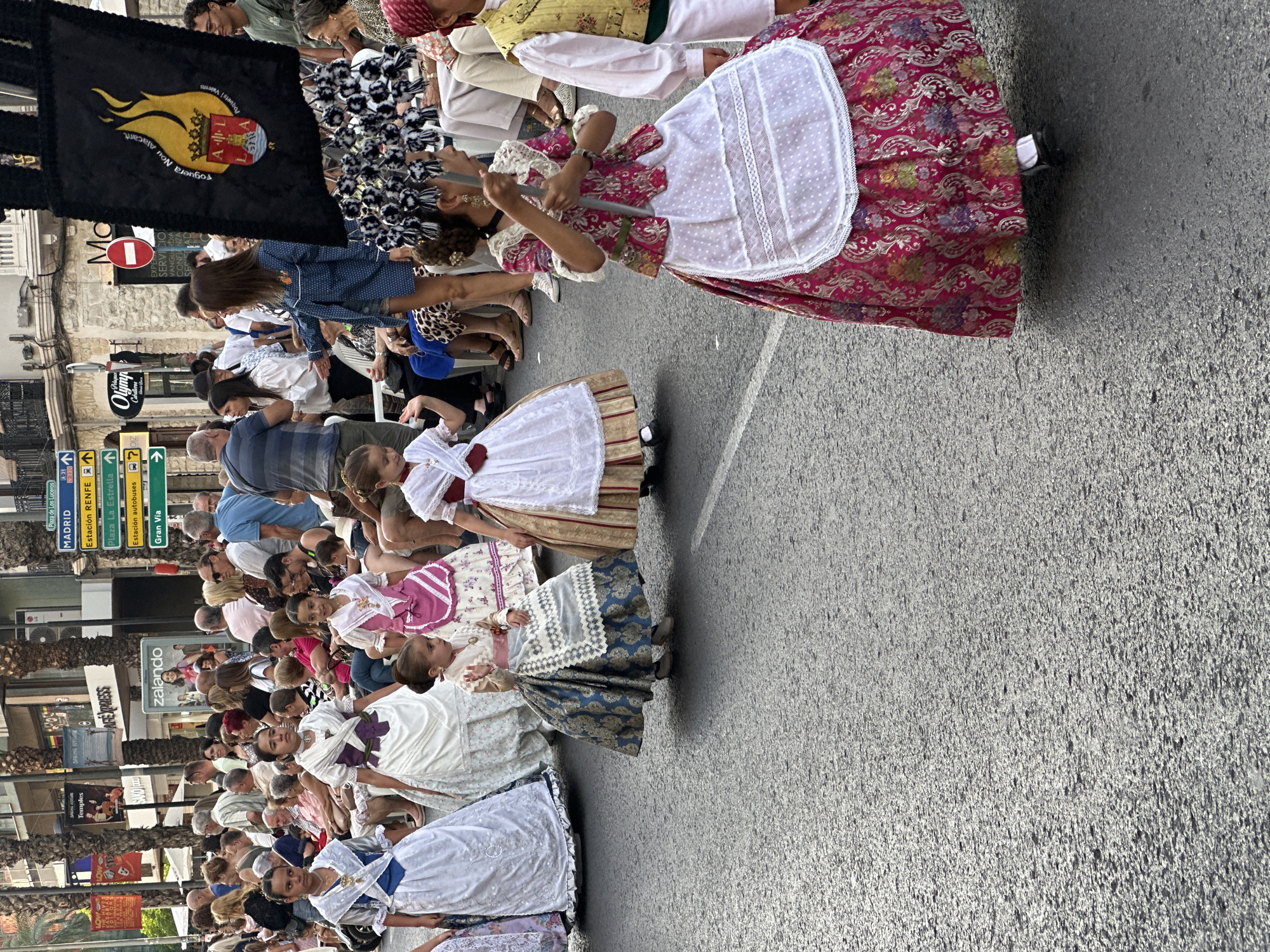 A street procession is taking place in Alicante, Spain. The street is paved with asphalt, and there are palm trees lining the sidewalk, suggesting a warm climate. Several girls in traditional Valencian dresses are walking in the procession. These dresses are elaborate, featuring full skirts with intricate patterns and lace detailing. Some of the girls are holding onto a banner with a flame emblem. Spectators are seated on chairs on the sidewalk, watching the event. Various buildings with shops and signs are visible in the background, including "zalando" and street signs indicating directions to "MADRID" and "Plaza de Los Luceros." The overall atmosphere appears festive.