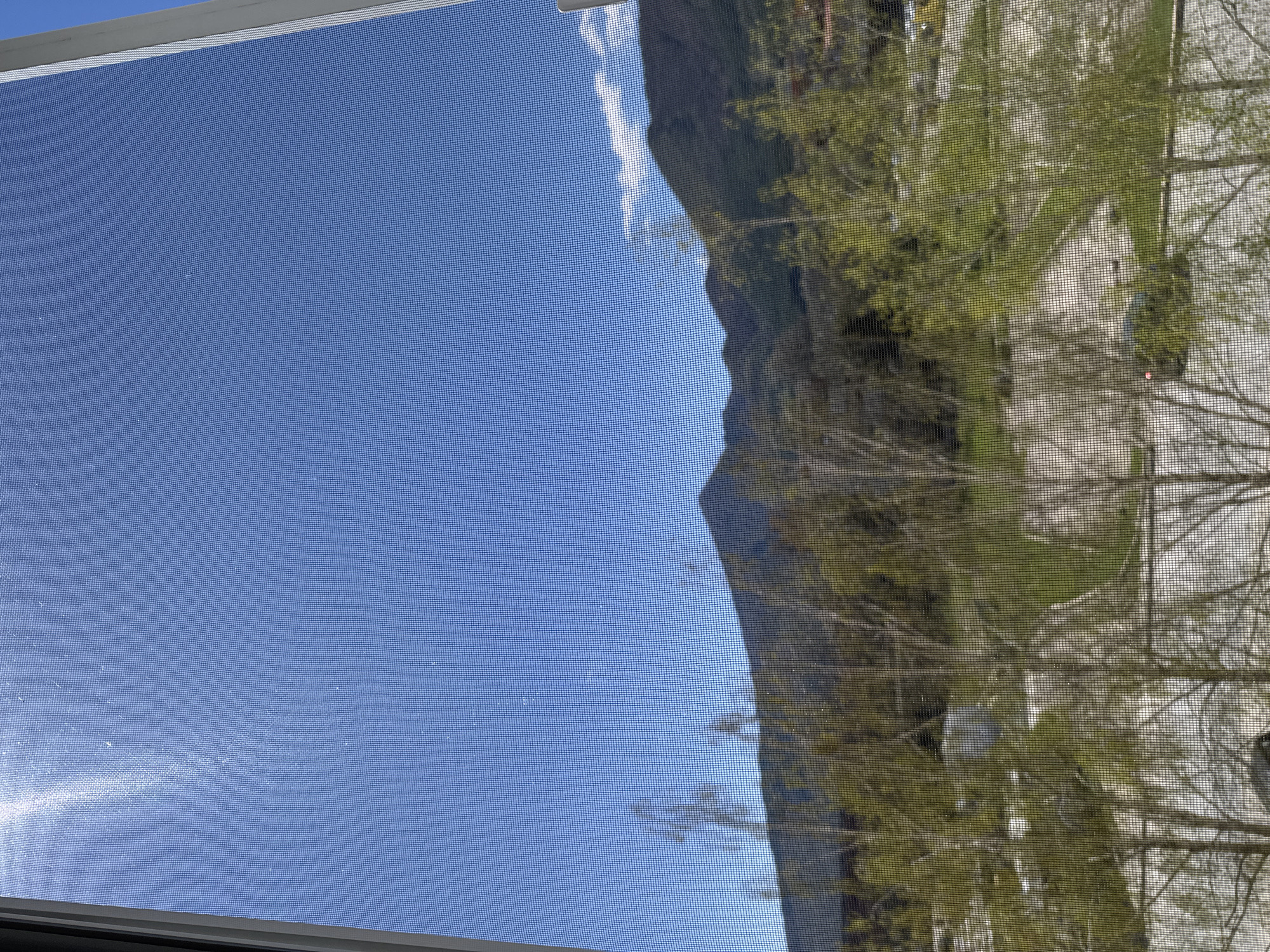 The image, taken through a window screen, shows a bright blue sky on the left side. On the right, the reflection in the glass captures a mountain range, vegetation (possibly trees), and a building. The mountain range suggests a location in a valley or near mountainous terrain. Without more specific architectural details or landmarks, it's not possible to confirm the location within Goce Delcev, Bulgaria.