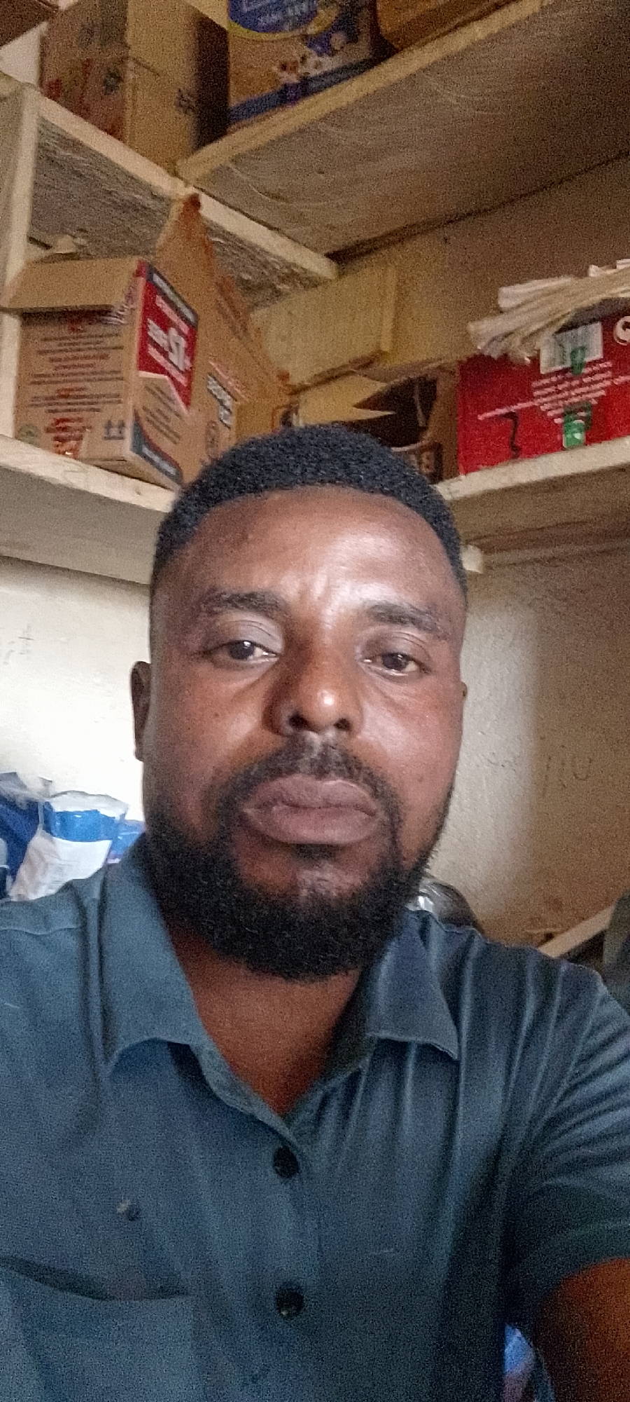 A close-up, waist-up, selfie-style photograph shows a Black man with short, dark, curly hair and a full beard and mustache. He is wearing a dark blue collared shirt with two black buttons visible at the neck. The man's face is turned slightly towards the camera, and he is looking directly at the lens. His eyes are open, and his lips are slightly parted.
The background of the image consists of shelves filled with various items, suggesting a storage area or a shop. On the upper shelves, there are boxes and packages, some with red and white labeling. On the left side, there are blue and white packaged items. The lighting appears to be artificial and somewhat dim, casting a slight shadow on the man's face. The overall setting is indoors. The image does not provide any specific indicators to determine its precise location within Durumi, Nigeria.