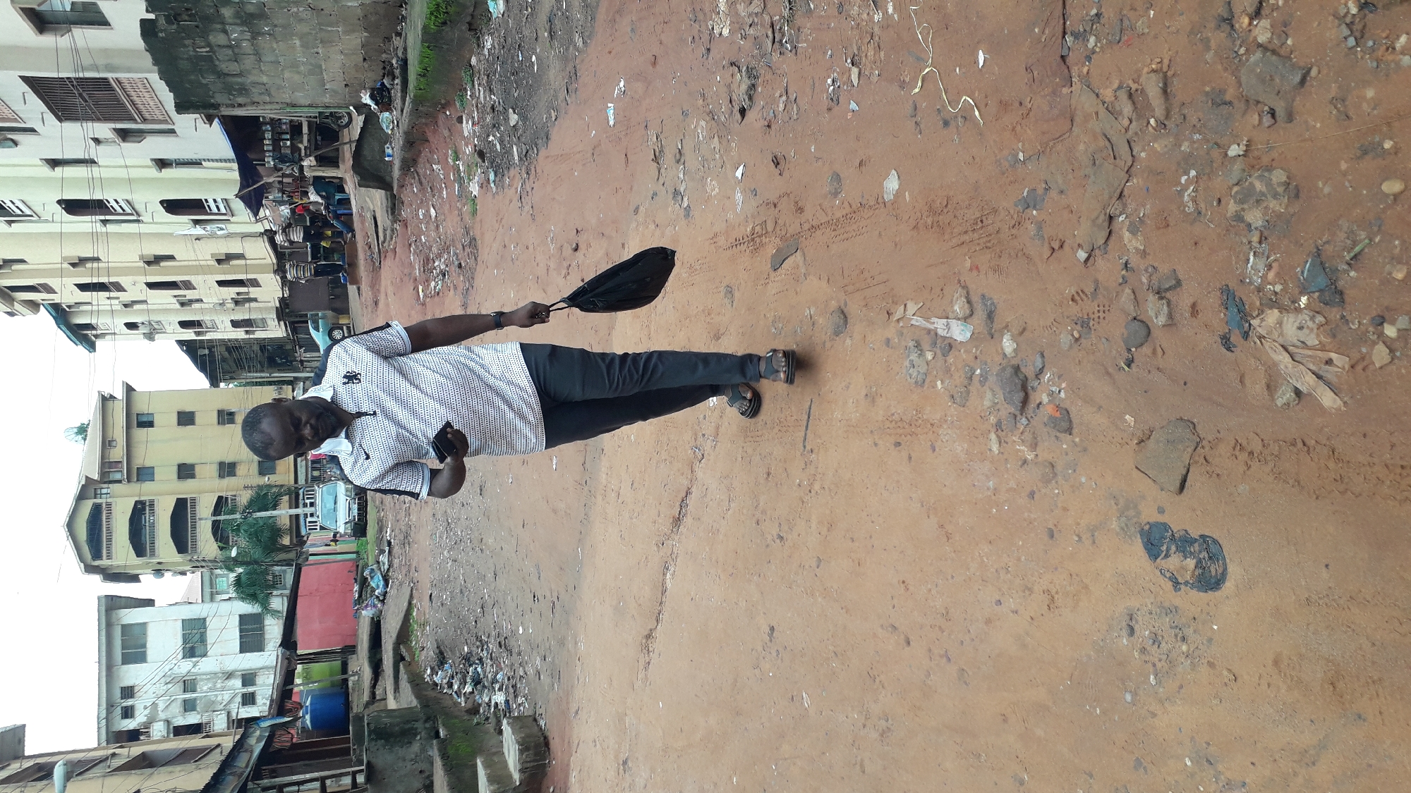 A man stands on a dirt road in Onitsha, Nigeria. He is wearing a patterned white shirt, black pants, and sandals. He holds a cell phone in his left hand and a black bag in his right. The road is covered in dirt, rocks, and trash. Buildings are visible in the background. There is garbage lining the sides of the road.