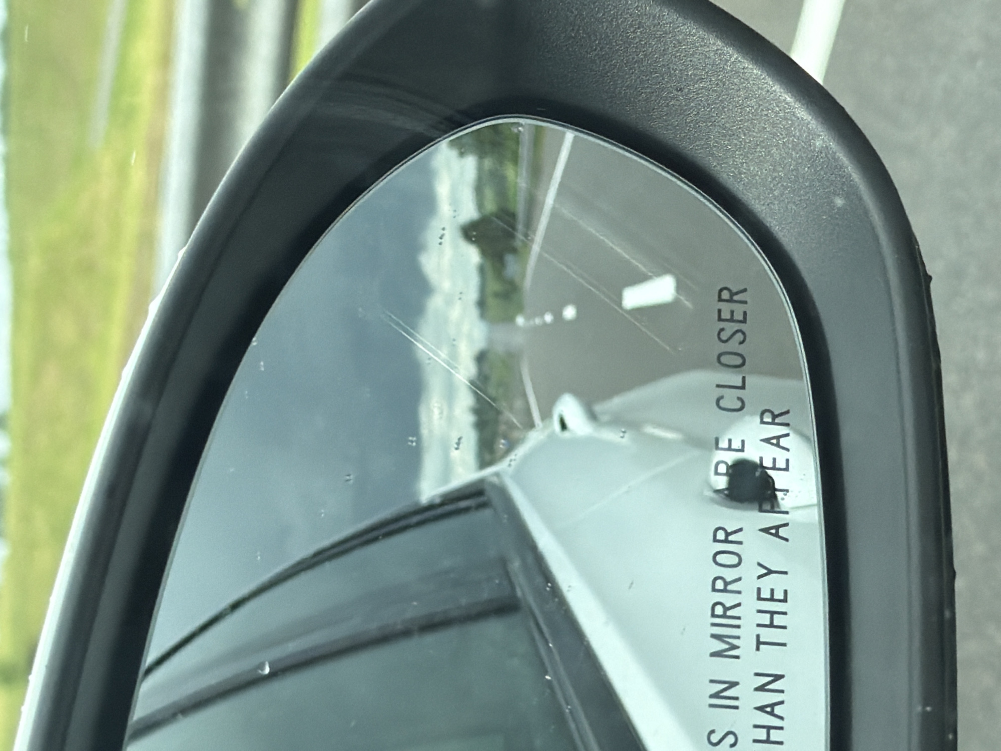 The image shows a close-up of a car's side-view mirror, reflecting a highway scene. The mirror displays text that reads "OBJECTS IN MIRROR ARE CLOSER THAN THEY APPEAR". The reflection shows a road with white lane markings, green fields, and trees on either side. The sky is cloudy. The mirror itself has some water droplets and fine scratches on its surface. The exterior of the car, a silver sedan, is visible on the left side of the image. The location is specified as Пяршайскі сельскі Савет, Belarus. There are no people visible in the image, and no actions or interactions are taking place. The environment appears to be rural, with a focus on the road and surrounding landscape as seen through the mirror.