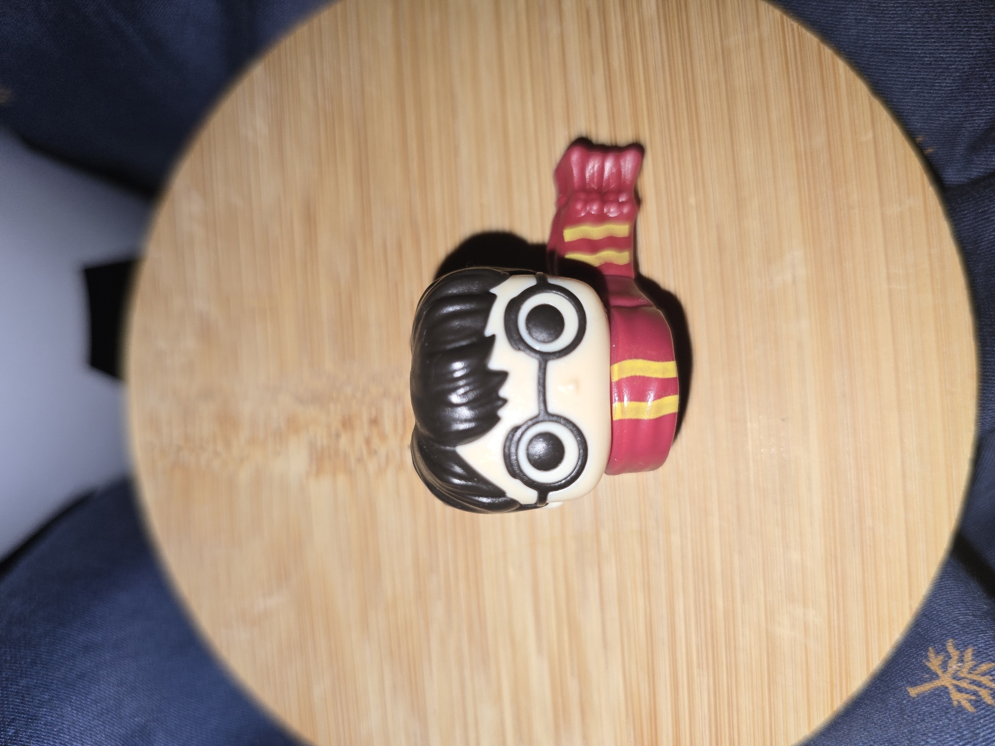 A close-up, overhead shot shows a Harry Potter Funko Pop figurine with a red and yellow striped scarf, resting on a round wooden surface. The wooden surface has a visible grain pattern and is slightly textured. The background consists of dark blue fabric, possibly clothing, with a faint pattern visible on the lower left side of the image. The lighting creates a soft shadow to the right of the figurine. This scene is not located in Petropavlivska Borshchahivka, Ukraine.