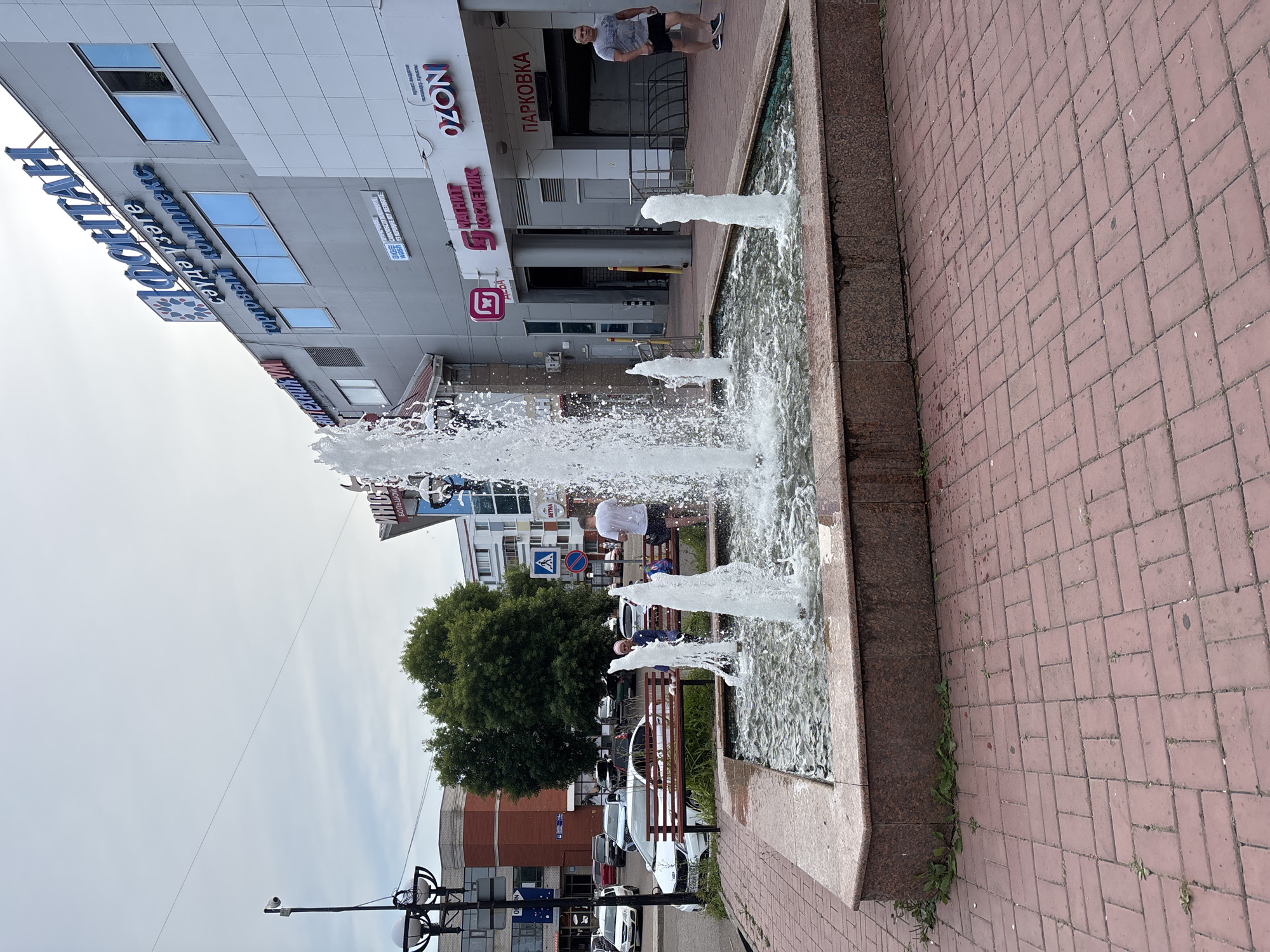This image captures a public scene in the city of Naberezhnye Chelny, Russia, featuring a prominent fountain in the foreground. The fountain has a square, stone basin with reddish-brown granite detailing and several water jets actively spraying water. The surrounding area is paved with reddish brick pavers arranged in a herringbone pattern.
Behind the fountain, a white vehicle is parked on the left side of the frame, and a red brick building with a white trim is visible. Several trees with lush green foliage are present, some obscuring parts of the buildings.
Dominating the background is a modern, multi-story building with a grey facade. It features large blue-tinted windows and several signs with Russian text. Notably, one large blue sign reads "ФОНТАН" (Fontan), indicating the name of the location or a business. Other signs are visible, including "OZON" and "ПАРКОВКА" (Parking).
Several people are present in the scene. On the right side of the fountain, a man in a grey t-shirt and black shorts walks away from the camera. To the left of the fountain, two individuals are seated on a wooden bench. One person, wearing a blue shirt and headscarf, appears to be looking towards the fountain. Another person, partially obscured, is also on the bench. Further in the background, near the building entrance, more people can be seen, some walking and others standing. The sky above is overcast with a light grey hue. The overall scene depicts an urban public space with elements of commerce and recreation.