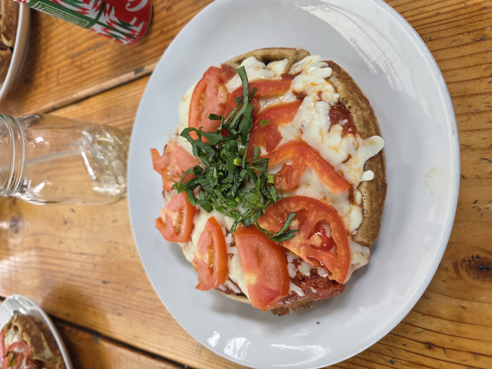 A high-angle, close-up shot presents a plate of waffle-based pizza, topped with tomato slices, mozzarella cheese, and fresh basil, on a white plate. The plate rests on a wooden table. To the left, a partially visible Mason jar and a red and green can, presumably a soft drink, are also on the table. Another plate with a similar dish is visible in the lower-left corner. The location is Ensenada, Mexico.