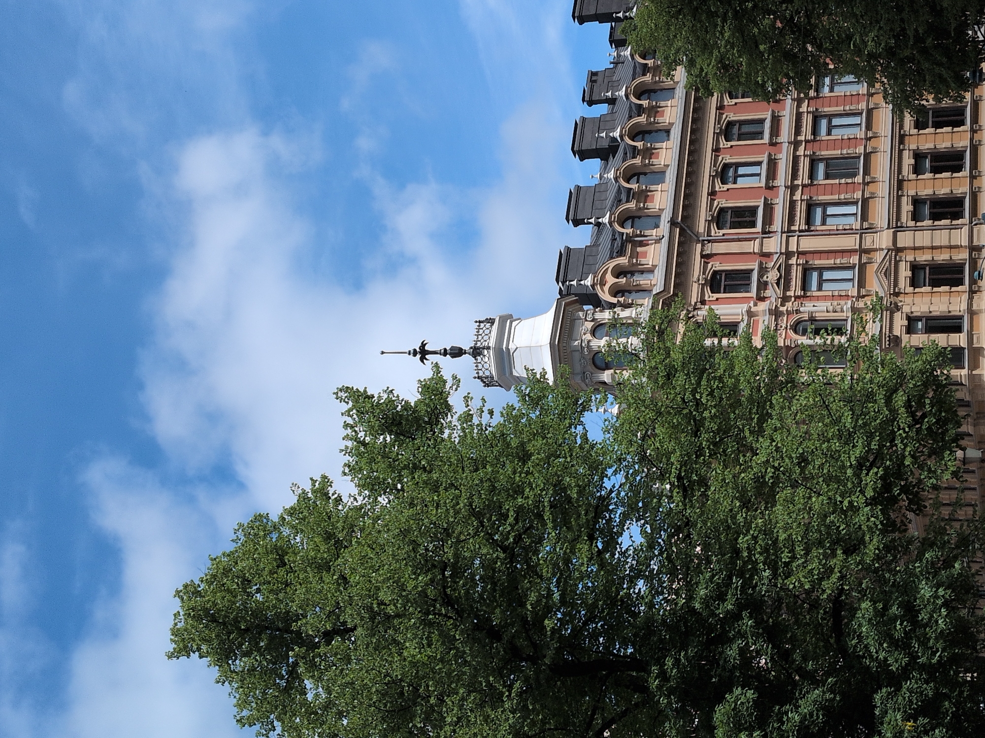 A building with ornate architecture, featuring a reddish-orange facade with yellow trim and arched windows, is partially visible through lush green trees under a bright blue sky with scattered white clouds. The building has a distinctive white tower topped with a decorative spire. This view is from the perspective of Esplanadi Park in Helsinki, Finland, on a clear day.
