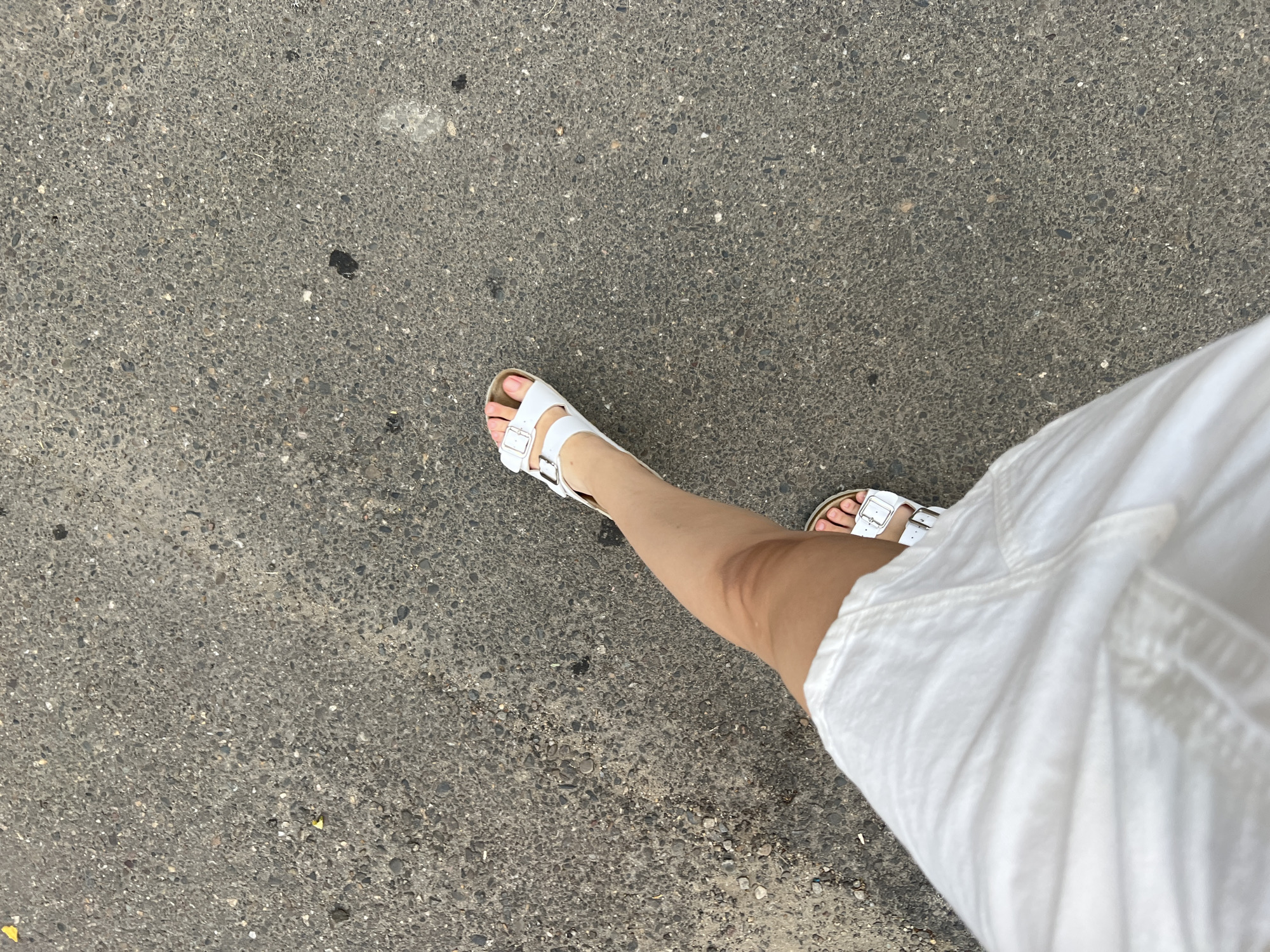 A person's legs and feet are shown from a top-down perspective, walking on a textured gray asphalt surface. The person is wearing a white dress that extends into the lower left corner of the frame, and white Birkenstock-style sandals on both feet. The asphalt has a rough, pebbly texture with scattered small debris. The scene is outdoors, likely in a city, but the specific location in Belozernyi, Russia, cannot be determined from the visual information alone. The lighting suggests it is daytime.