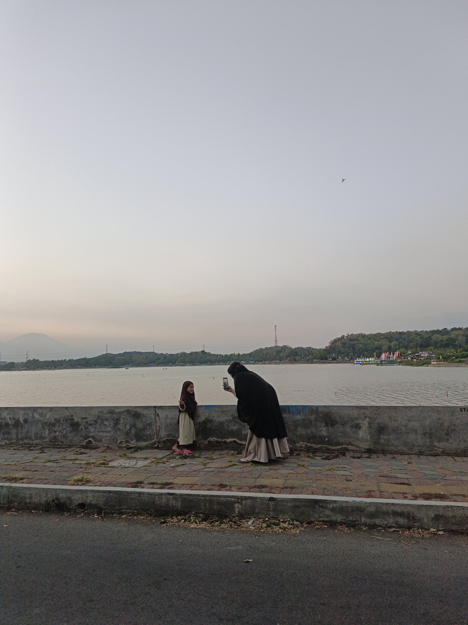 Two individuals, a woman in a black hijab and a long skirt, and a young girl in a brown hijab and a beige dress, stand on a paved walkway beside a body of water. The woman is holding a smartphone, seemingly taking a picture of the girl, who is facing away from the camera and looking to the side with her mouth slightly open. The walkway is bordered by a concrete wall that separates it from the water, and behind the wall, a calm body of water stretches towards a treed shoreline. In the distance, hills and mountains are visible under a pale, overcast sky. The scene appears to be an outdoor public space, possibly a park or a promenade, near Krakitan, Indonesia.