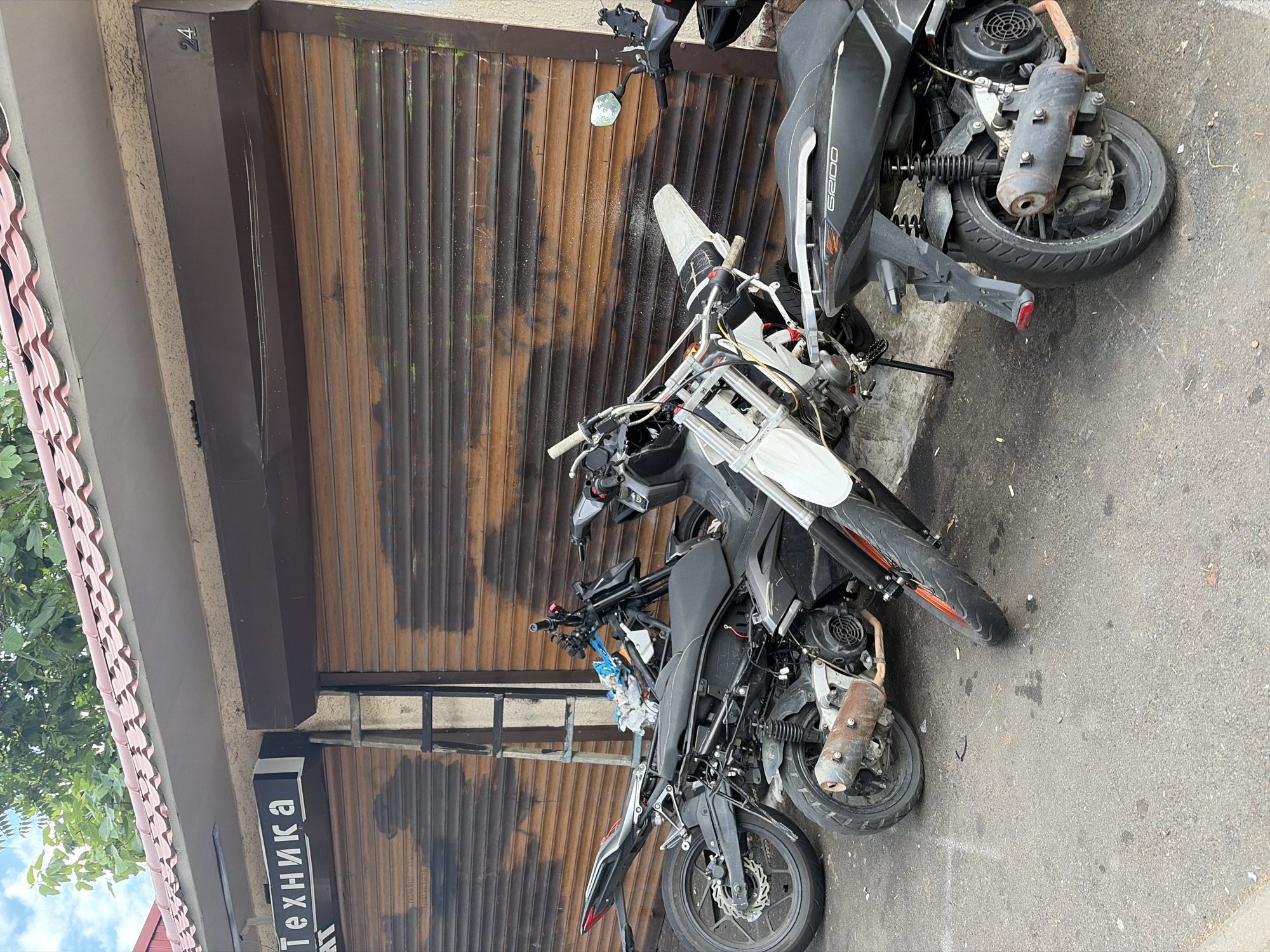 A street scene in Sochi, Russia, shows three parked motorcycles and scooters in front of a building with a corrugated metal roll-up door. A sign above the door reads "Техника" in Cyrillic letters, meaning "Technics" or "Equipment." The motorcycles appear to be a mix of dirt bikes and scooters, some in better condition than others. One dirt bike in the foreground is partially disassembled, with its front end propped up. The ground is asphalt, with scattered debris. The building itself has a tiled roof and a tan-colored facade. The lighting suggests it is daytime, with natural light illuminating the scene.