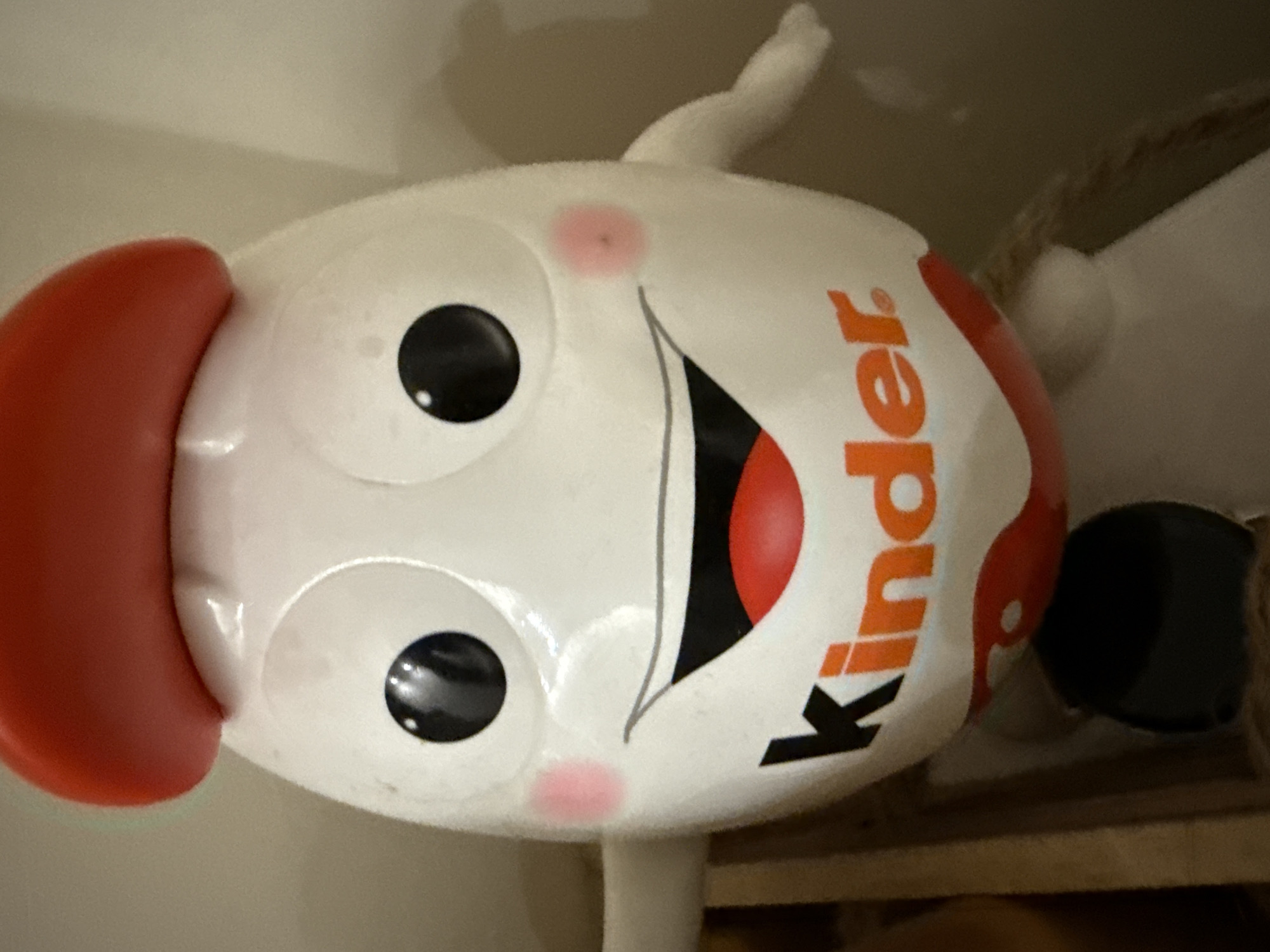 The image features a cheerful, egg-shaped character, likely a mascot for the Kinder brand, crafted from a white, soft-looking material. The character has a large, red, semi-circular brim or hat on its left side. Its face displays two prominent black eyes with white pupils, two pink blush spots on its cheeks, and a wide, smiling mouth with a red interior and a black outline.
The word "Kinder" is visibly printed in orange letters, oriented vertically, on the right side of the character's face, accompanied by a red and white pattern. The object appears to be a plush toy or a decorative item, presented in an upright position.
The background consists of a light-colored, indistinct surface, possibly a wall. Beneath the character, a brown, horizontally patterned surface, resembling wood, is visible. Other soft, white appendages or parts of the toy are partially visible around the main figure. The character's expression is notably happy and inviting. The scene is static, showing no specific activity or event. The time of day and exact outdoor location cues are not discernible from the image.